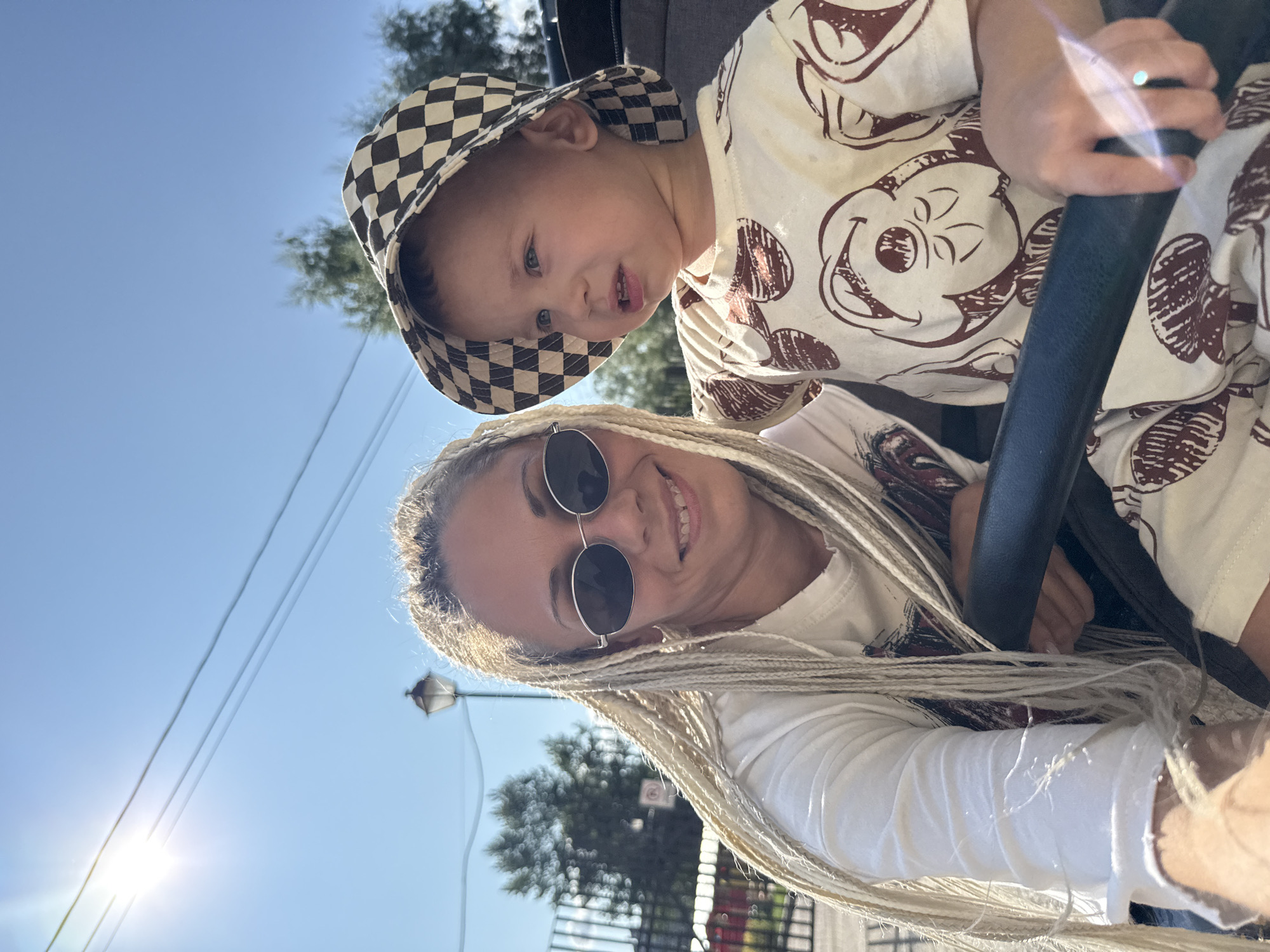 The image captures a woman and a young child, possibly mother and son, posing for a selfie outdoors under a bright, sunny sky. The woman has long, blonde braided hair and is wearing sunglasses and a white t-shirt. She is smiling at the camera. The child, wearing a checkered bucket hat and a light-colored t-shirt with a repeating pattern, is looking off to the side. They appear to be sitting together, possibly in a stroller or a similar seating arrangement, as suggested by the dark bar in the foreground. In the background, there is a black metal fence, trees, a lamppost, and power lines, indicating an outdoor urban or suburban setting. The sun is prominently visible in the upper left corner of the frame, casting a strong light. The location is identified as Rusanovo, Russia.