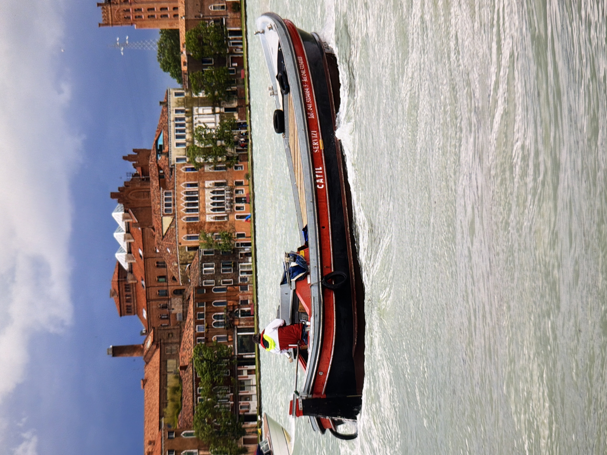 The image shows a red and black motorboat with "CATIL SERVIZI" and telephone numbers printed on its side, moving through choppy water. A person wearing a red and white jacket is seated at the helm. In the background, a dense collection of terracotta-roofed buildings with arched windows lines the waterfront, under a cloudy, dark blue sky. The buildings appear to be old and European in style. There is no indication that this scene is located in Kyiv, Ukraine.