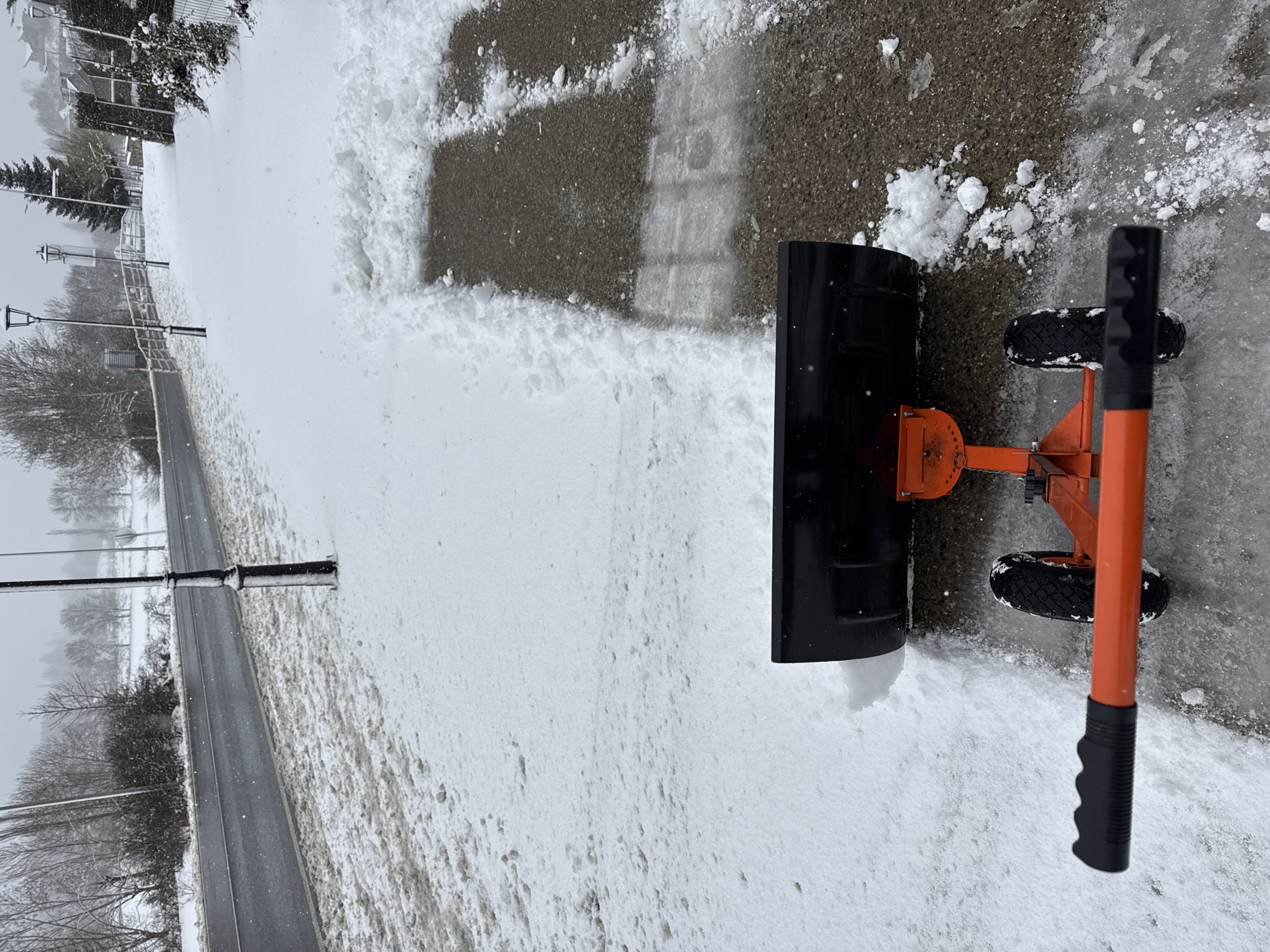 The image shows a winter scene with a snow pusher on a sidewalk. The snow pusher is orange with black handles and a black blade, and it is positioned on a sidewalk that has been partially cleared of snow. Snow is piled up on either side of the cleared path.
In the background, a road and buildings are visible. Bare trees and streetlights line the road, and the buildings are partially obscured by the snowy conditions.
The image does not provide enough information to determine if it is located in Benasque, Spain.