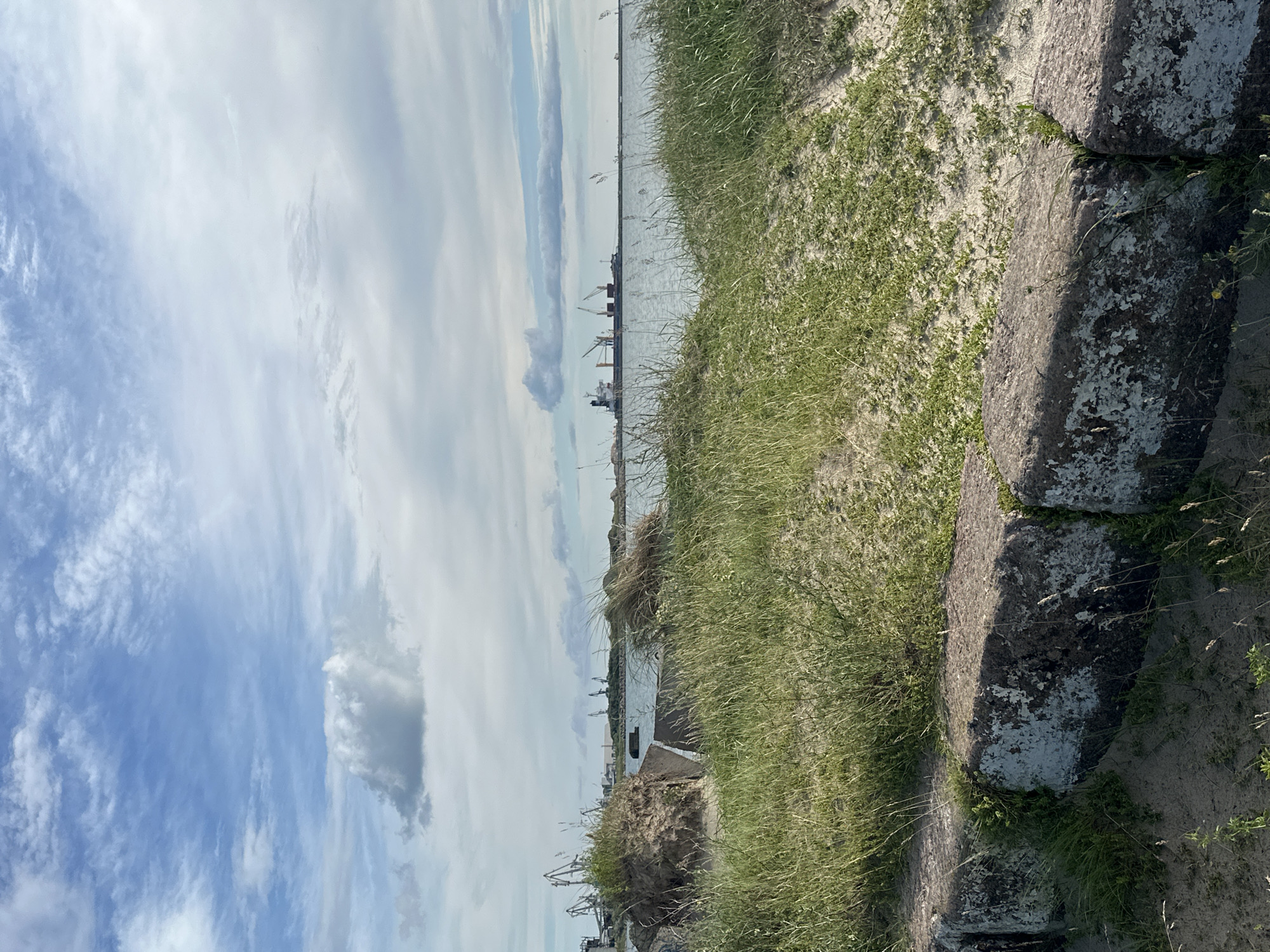 The image depicts a view of the Liepaja Port in Latvia on a cloudy day. In the foreground, there are large, weathered concrete blocks partially covered in sand and grass, suggesting a coastal or harbor edge. Beyond the blocks, there is tall, green grass and sandy terrain. The middle ground shows the calm sea water. In the background, the silhouette of the Liepaja Port is visible with several industrial cranes and ships docked or anchored. The sky is a mix of blue with scattered white and grey clouds, indicating a dynamic weather pattern. The overall scene is one of an industrial port area bordering a natural coastal landscape.