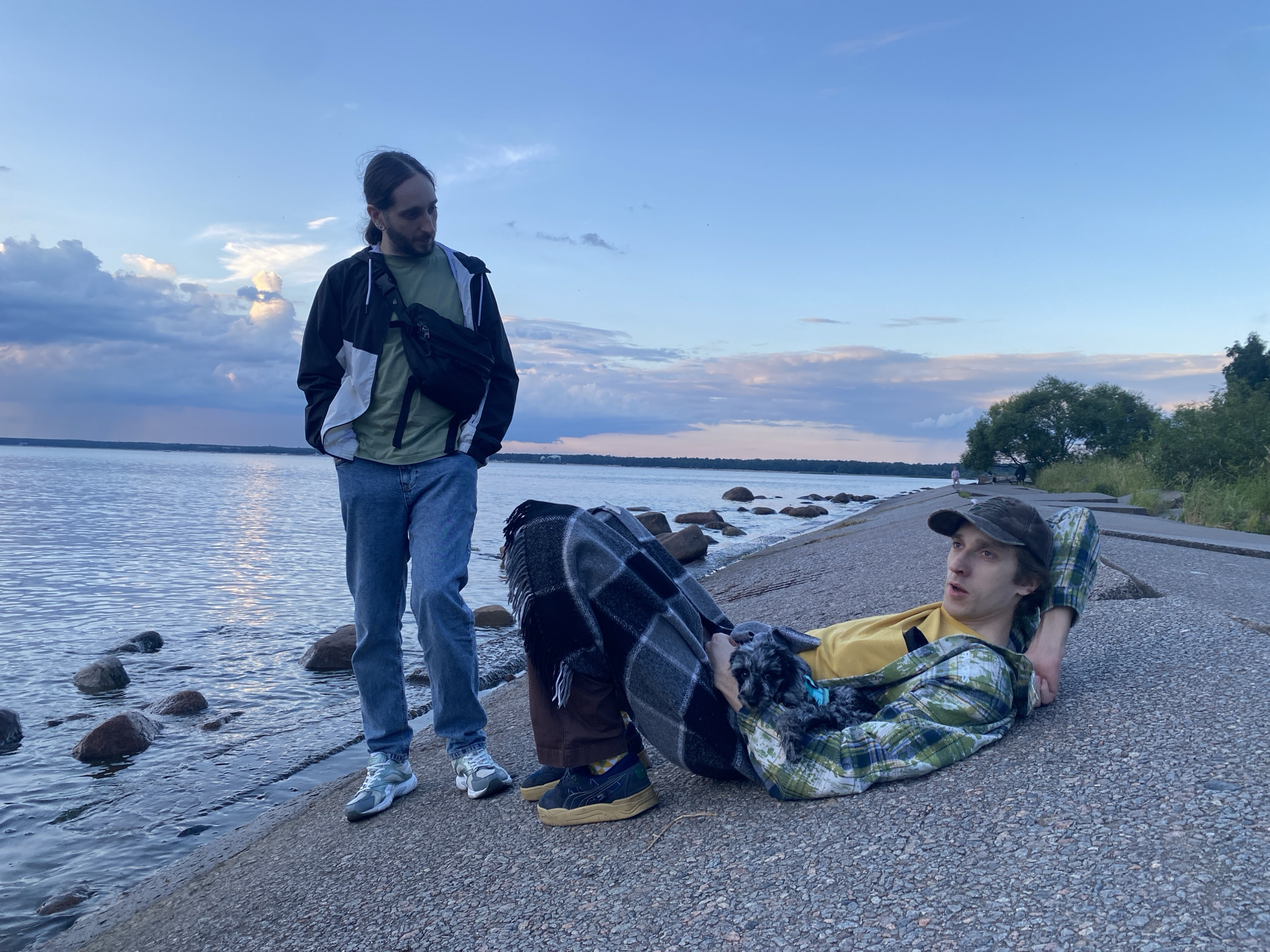 Two men are standing and lying on a concrete embankment next to a body of water, likely in Sestroretsk, Russia, given the context. The man standing on the left wears a black and white jacket over a green t-shirt and blue jeans. He has a dark fanny pack across his chest. The man lying on the right wears a yellow shirt and a green plaid jacket. He has a baseball cap on and is partially covered by a checkered blanket. A small, dark-colored dog is nestled on his lap, also partially covered by the blanket. The water is calm with some rocks visible near the shore. The sky is a dusky blue with scattered clouds, suggesting it is late evening. Trees and vegetation are visible in the background on the right side of the frame.