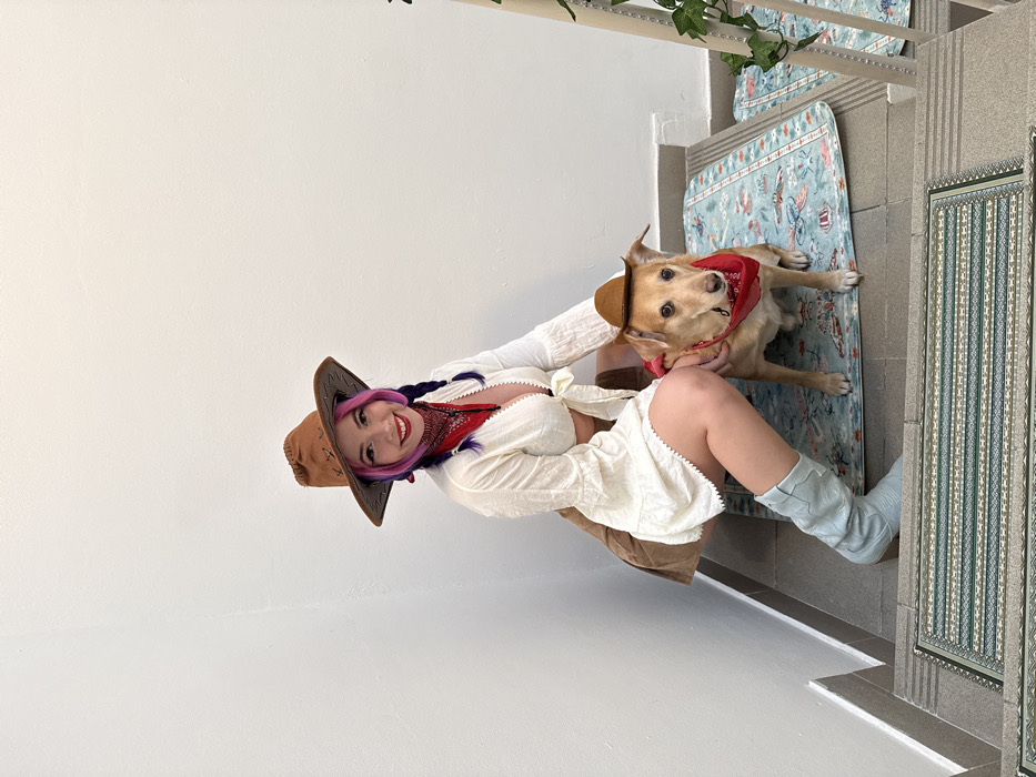 A woman and a dog, both wearing cowboy hats, are shown on a patterned rug. The woman is wearing a white, low-cut top, matching skirt, and light blue cowboy boots. The dog is also wearing a red bandana. The woman has purple hair with braids and is smiling. The background includes a white wall, a stairway with patterned rugs, and some greenery.