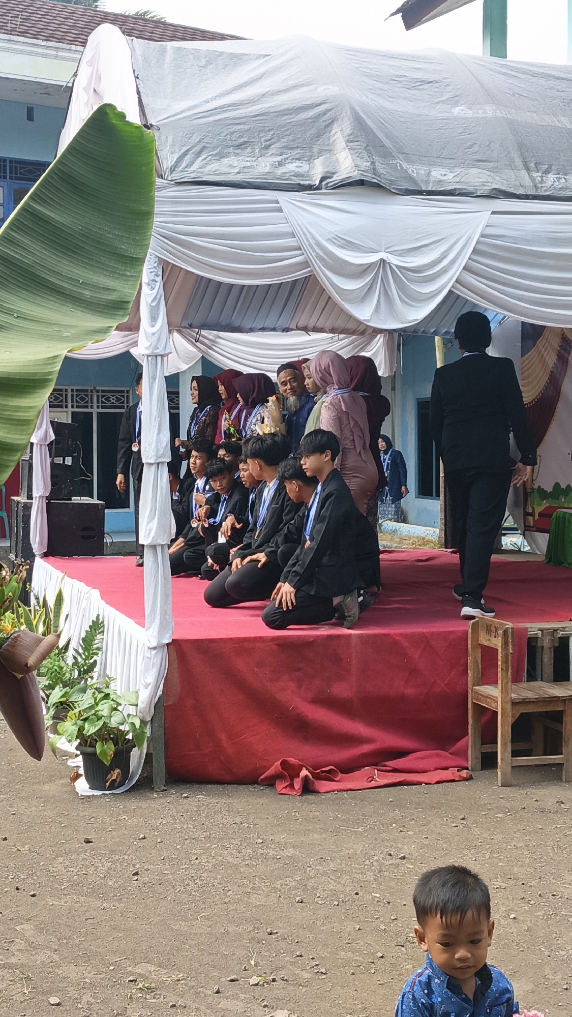 In front of a blue building in Tigaraksa, Indonesia, an outdoor event is underway. A stage is set up with a red carpet and draped white fabric, creating a ceremonial atmosphere. A group of young men in formal black suits and medals are kneeling on the stage, possibly receiving awards. Behind them, a line of adults, including women in headscarves and men in suits and a white beard, stand and observe. One man in a black suit stands facing away from the camera on the right side of the stage. In the foreground, a young boy in a blue patterned shirt looks down while holding something pink. The ground is dirt and gravel, with a large green leaf from a banana tree in the left foreground. The backdrop suggests a school or community event.