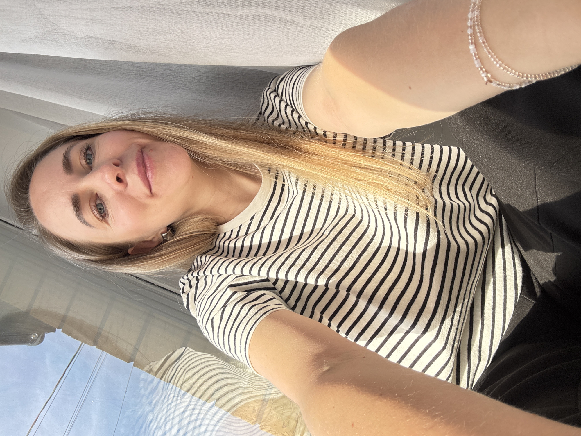 This brightly lit, candid selfie features a young woman with long, straight blonde hair, fair skin, and light-colored eyes, gently smiling directly at the camera. She appears to be indoors, near a window in Chaikovsky, Russia. She is wearing a light-colored (possibly white or off-white) t-shirt with black horizontal stripes and dark (likely black) pants or a skirt. A delicate, multi-strand beaded bracelet adorns her right wrist.
Strong natural light illuminates her from the left, casting a warm glow on her skin and hair, indicating daytime. The background shows a translucent white or light-colored curtain or blinds in the upper portion of the image. A clear reflection in the lower-left part of the window reveals a bright blue sky, overhead wires, and some distant building structures, confirming clear weather outside. The overall atmosphere is calm and pleasant. No text is visible in the image.