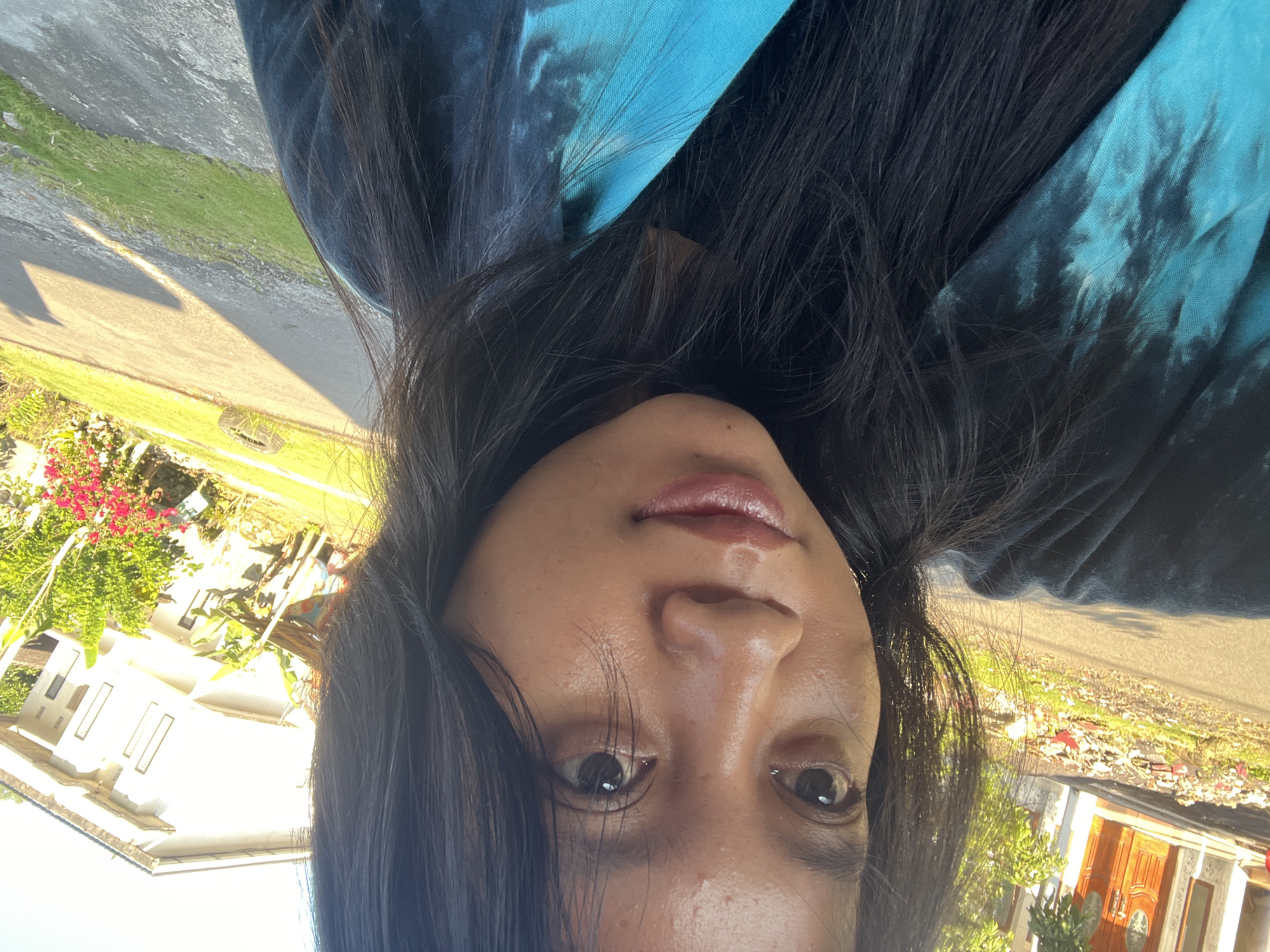The image is an outdoor selfie featuring an individual, likely female, with dark hair and a medium complexion, taken in Unknown, Unknown. She is positioned for a close-up, with her gaze directed slightly away from the camera. Her hair is dark, straight, and frames her face. She wears a black garment beneath a blue and black tie-dye top or wrap. The background depicts a residential street or pathway, partially paved, with patches of green grass and various plants. To the left, a house with a light-colored exterior and a prominent brown double door with decorative glass is visible. To the right, another residential structure with a white or light-toned facade and multiple rectangular windows can be seen. Foliage and plants with red flowers are present along the roadside. Strong natural sunlight illuminates the scene, casting distinct shadows on the ground, indicative of daylight conditions.