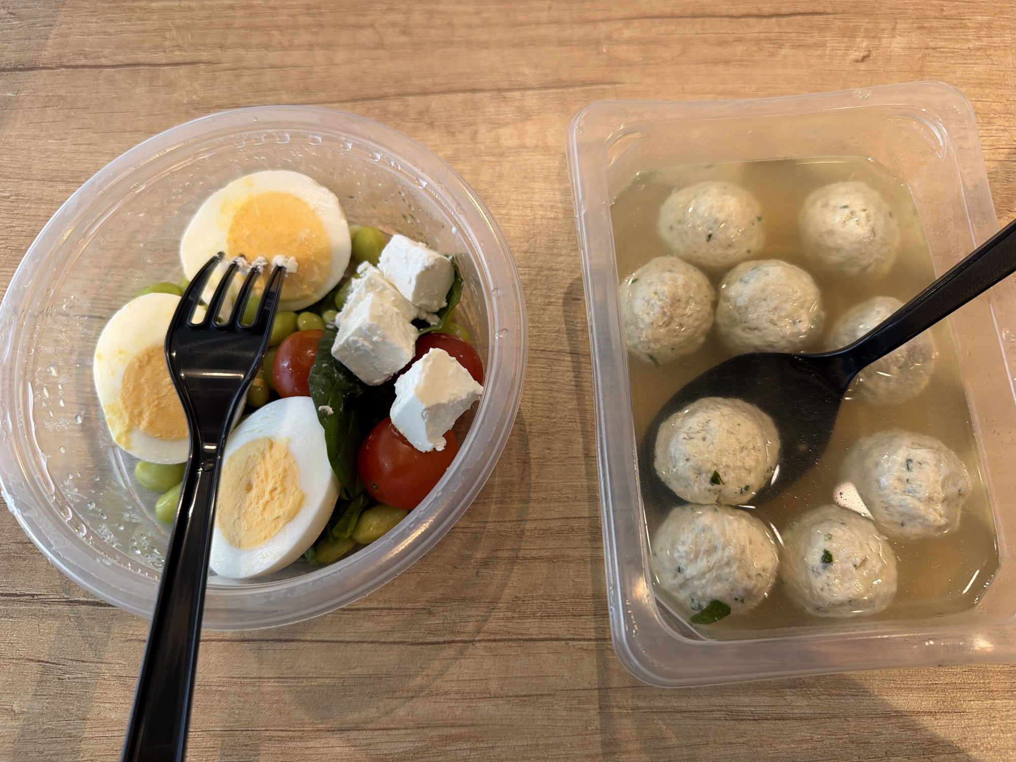 The image displays two clear plastic food containers set on a light-colored wooden table, suggesting an indoor setting such as an office, home, or cafeteria in Moscow, Russia. The table surface features a visible wood grain pattern.
The container on the left is round and holds a fresh salad. Key ingredients include several halves of hard-boiled eggs with firm, bright yellow yolks, small green edamame beans, and vibrant red cherry tomatoes. Cubes of white, soft cheese, likely feta, are scattered generously across the top, and some green leafy vegetables, possibly spinach, are visible beneath the other components. A black plastic fork is positioned within this salad, indicating it's ready for consumption.
The rectangular container on the right holds a light-colored, clear broth or soup containing approximately ten pale, spherical items. These items resemble chicken or fish meatballs or dumplings, with subtle flecks of green herbs visible on their surface. A black plastic spoon is partially submerged in the broth among these floating components.
The overall impression is a prepared, convenient meal, likely a lunch, packaged for takeout or easy transport. No people or visible text are present in the image.