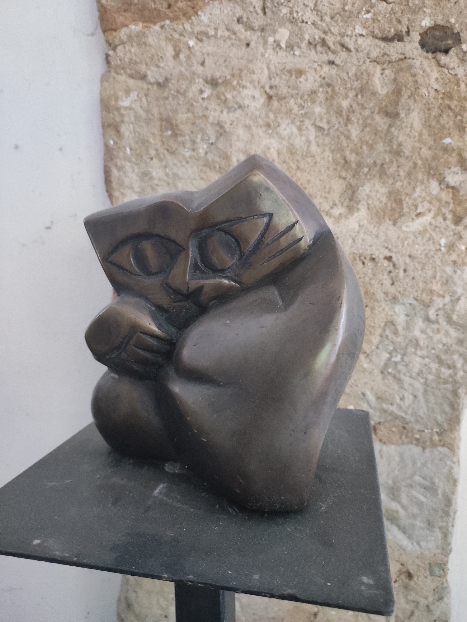 A bronze sculpture of a cat, depicted in a stylized, cubist manner, sits on a black square pedestal. The cat's face features angular, almond-shaped eyes with etched pupils, a small, triangular nose, and lines suggesting whiskers. Its body is rounded and folded, with one paw resting on its chest. The sculpture has a dark, aged patina with some lighter highlights reflecting the ambient light.
The sculpture is positioned in front of a rough, textured stone wall, which appears to be part of an older building. To the left of the sculpture, a plain, light-colored wall contrasts with the stone. The overall environment suggests an outdoor or semi-outdoor setting, possibly a courtyard or garden area within the city of Dresden, Germany. The lighting is diffused, indicating it is likely daytime. The image is a medium shot, focusing on the sculpture and its immediate surroundings.