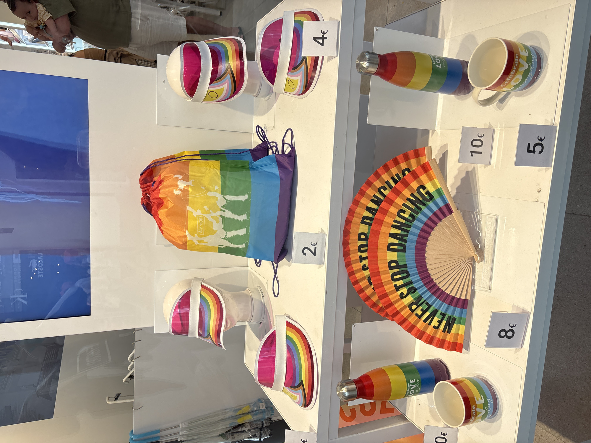 This is an indoor product display of rainbow-themed merchandise, likely a retail store or market stall. The items are arranged on white shelves. In the foreground, there are two rainbow-colored fans, a rainbow-patterned water bottle, and a rainbow-patterned mug. A price tag of "8€" is visible near the mug. Further back, a larger rainbow drawstring bag with graphics is displayed. To the left and right of the bag are rainbow-patterned visors on mannequins, with price tags of "2€" and "4€" respectively. On the far left, partially visible, are umbrellas with blue handles. In the background, a large screen displays text and imagery, and a person is holding a baby. The overall aesthetic suggests items related to LGBTQ+ pride. There is no visible indication of the city of Orihuela, Spain, within the image or the provided crops.