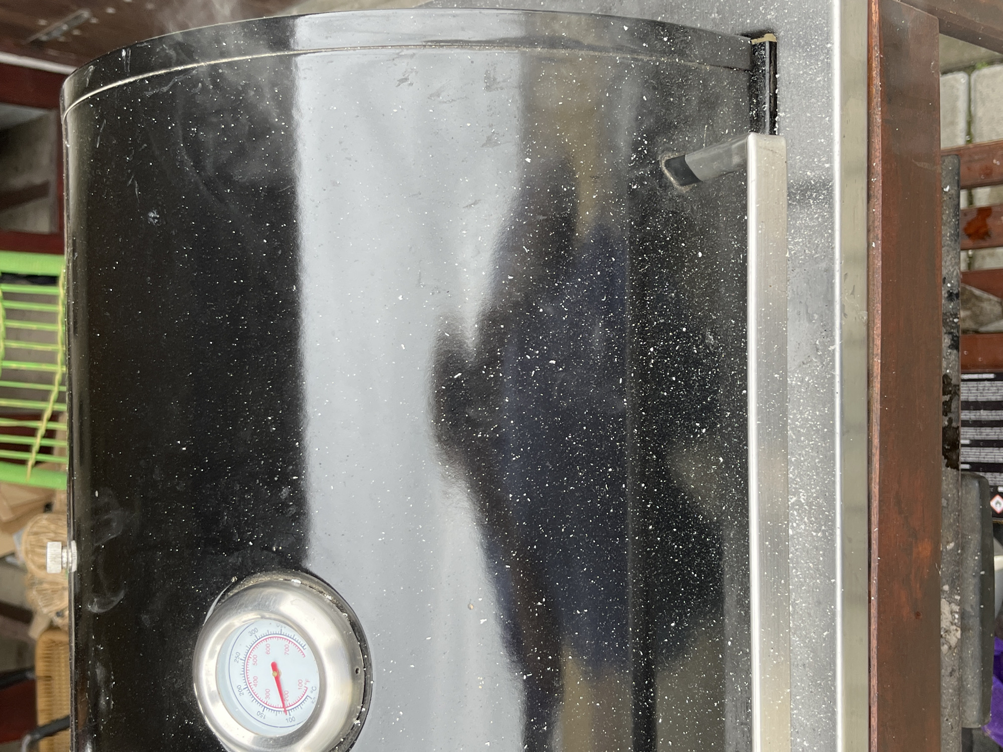 This image shows a close-up of a black outdoor smoker or grill. The smoker features a built-in thermometer with a red needle, located on the lower front. The thermometer face has temperature markings in degrees, indicated by the printed text. The smoker's surface appears speckled with white spots. There is a metallic handle and trim on the right side of the smoker. Smoke or steam is visible emanating from the top edge of the smoker, suggesting it is in use or recently used. A partial view of what seems to be wooden and metal patio furniture can be seen in the periphery. The environment suggests an outdoor patio or backyard setting. The location is unspecified but if it is in Pchelnik, Bulgaria as specified, no specific characteristics confirm this locality based solely on the image.