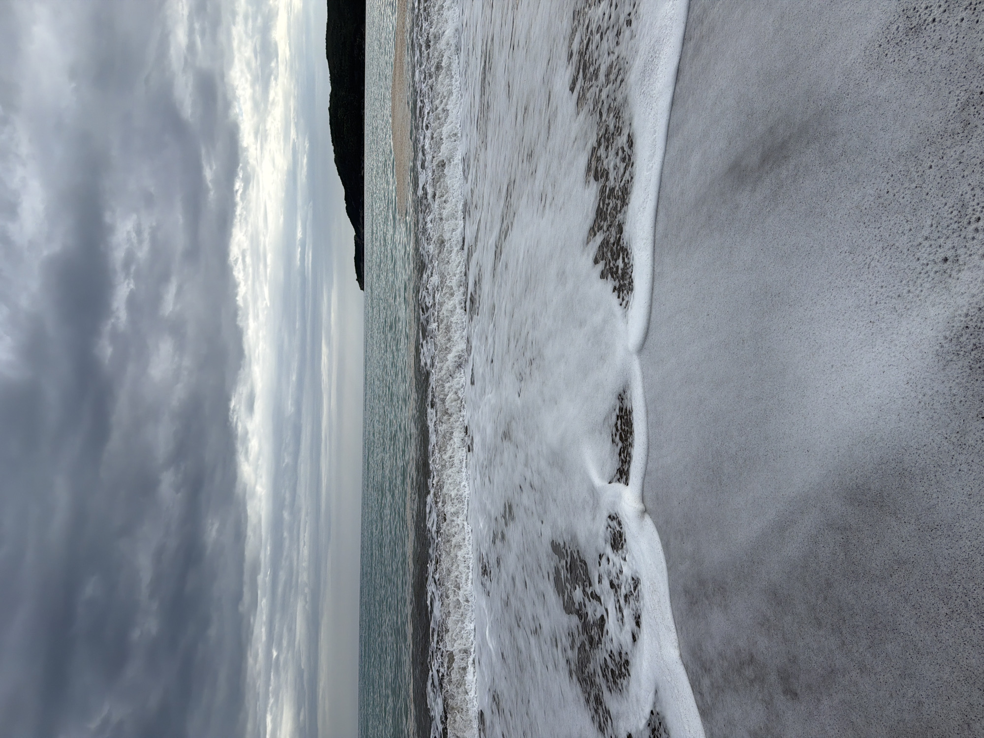 The image depicts a beach scene in Boreti, Montenegro under a cloudy sky. Waves are crashing on the shore, creating white foam. The sand is dark. A headland is visible in the distance. No people are present.