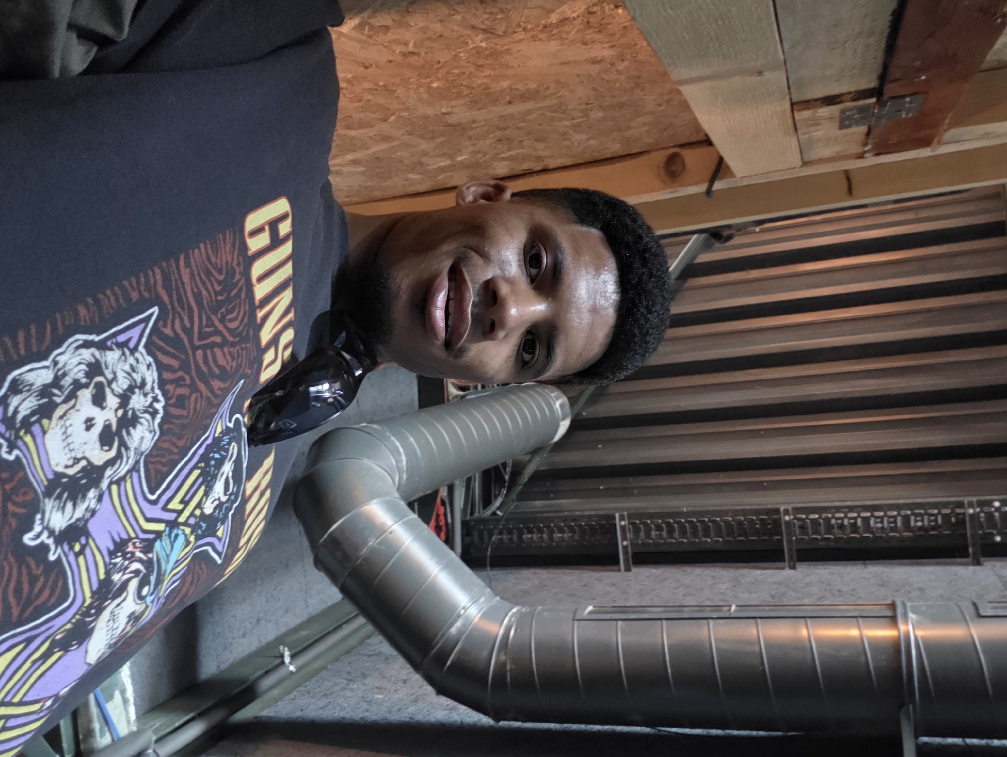 A young Black man is pictured in a close-up selfie, looking directly at the camera. He is wearing a dark t-shirt with a graphic design featuring skulls and the text "GUNS". He has short, dark, curly hair and a light stubble. He is smiling broadly, with his mouth slightly open. Behind him, the background consists of a light-colored metal duct system, a wall with vertical paneling, and wooden planks. The image appears to be taken indoors, possibly in a backstage area or industrial setting.