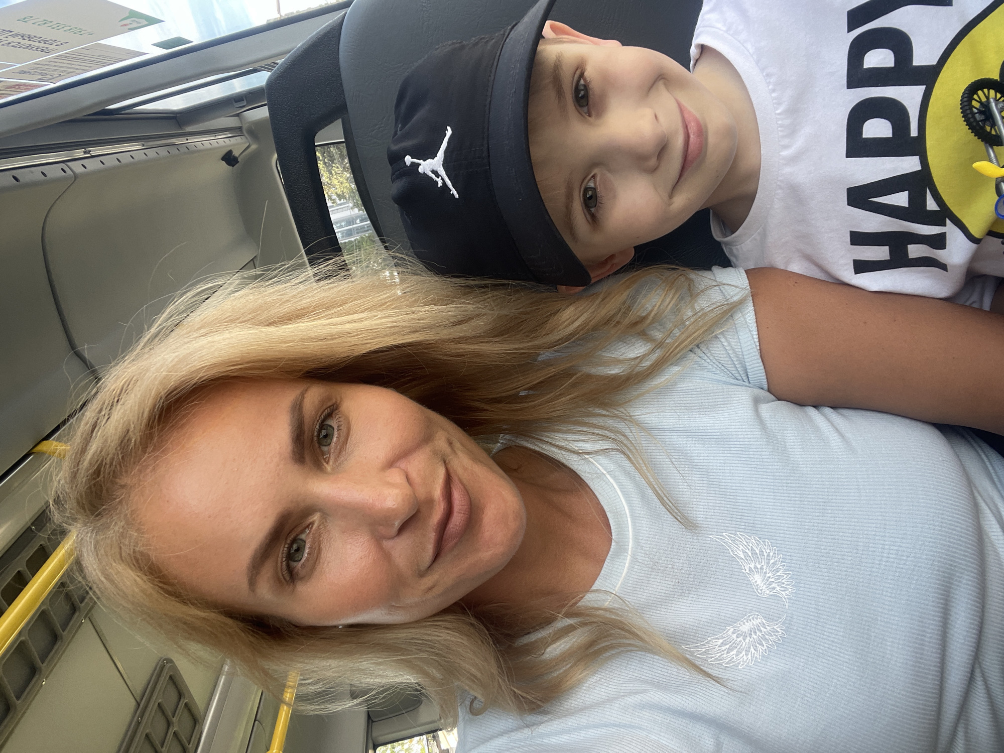 A joyful selfie captures a woman and a young boy, likely her son, inside a public transport vehicle in Sochi, Russia. The woman, with long blonde hair, is smiling gently, wearing a light blue ribbed shirt adorned with white winged embroidery. Beside her, a young boy with light brown eyes also smiles, wearing a black baseball cap featuring a white Jordan "Jumpman" logo and a white t-shirt. His shirt prominently displays the word "HAPPY" in black block letters, accompanied by a yellow smiley face holding a small bicycle model.
The setting appears to be the interior of a bus or similar vehicle, with gray overhead panels, dark-colored seats (a headrest is visible), and bright yellow grab rails. Bright daylight streams in from the windows or sunroof, indicating it is daytime with sunny or clear weather outside. Through the upper window, a white sign with partially visible text, possibly in Cyrillic, and numbers like "8/73 723 05-24" can be seen. Both subjects appear happy and content, sharing a pleasant moment during their journey.