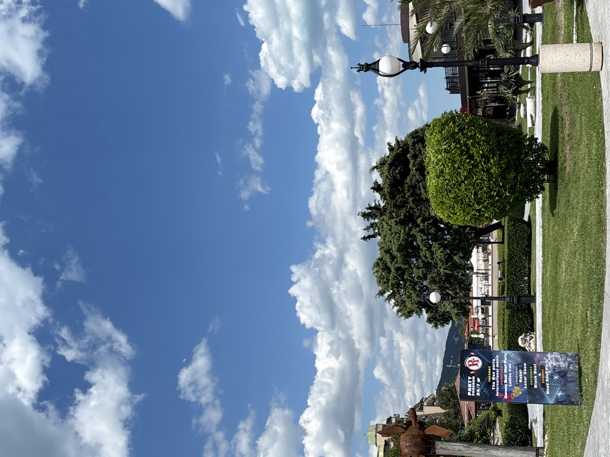 The image depicts a park scene in Varna, Bulgaria, with a clear blue sky dotted with white, fluffy clouds. In the foreground, a green grassy lawn is bordered by a paved walkway. To the left, a tall, dark sign displays a "PARTY PROGRAM 24/7" with listings for different days and events, including "The Bear Party International music," "Gummy Bear Shot Party," "Ladies free," "Robot Laser Show," "Black Beat," and "Head Shot Party." A large, ornamental, rusty metal sculpture is visible behind the sign. A neatly trimmed, spherical green bush stands in the center of the lawn. To the right, a black lamppost with a white globe stands near a cylindrical stone base. Beyond the lawn, a line of meticulously maintained hedges leads to trees and buildings in the background. Among the trees, the top of a building with a green dome is visible. Palm trees and other foliage suggest a warm climate. A small white object, possibly a kite or a bird, is visible in the sky, high above the landscape. Several white umbrellas and outdoor seating arrangements are present in the mid-ground, indicating a seating area or an outdoor cafe.