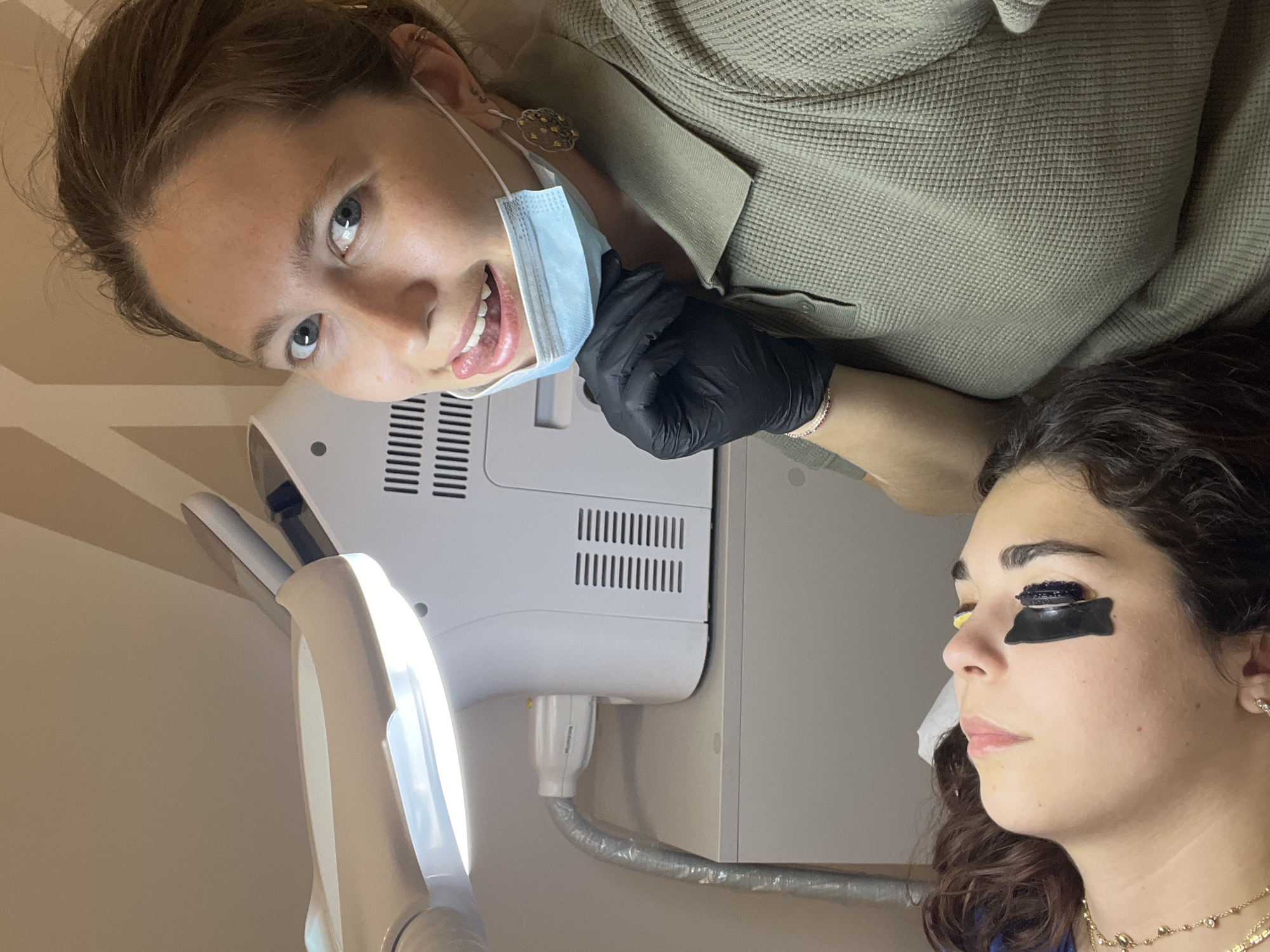 This image captures a beauty treatment session in an indoor setting, likely a salon or clinic in Podgorica, Montenegro. Two women are the primary subjects.
One woman, likely the esthetician, is positioned above and to the left, smiling and looking directly at the camera. She has light brown hair, blue eyes, and is wearing a sage green waffle-knit top. A light blue surgical mask is pulled down below her chin, revealing her smile, and she is wearing black disposable gloves. Earrings are visible on her left ear.
The second woman, the client, is lying down on the right side of the frame, seen in profile. She has dark, curly hair and is wearing multiple layered gold necklaces. Her eyes are covered with distinct black and yellow patches, indicating an ongoing beauty procedure, most likely an eyelash treatment. Her expression appears calm and relaxed.
In the background, behind the esthetician, white professional equipment is visible. This includes a machine with horizontal ventilation grilles and a prominent, curved white lamp arm that extends towards the client's face, providing bright illumination. Some indiscernible text is faintly visible on the side of the white equipment. The lighting suggests a well-lit indoor environment, possibly during the day. The esthetician's friendly expression contributes to a positive and professional atmosphere.