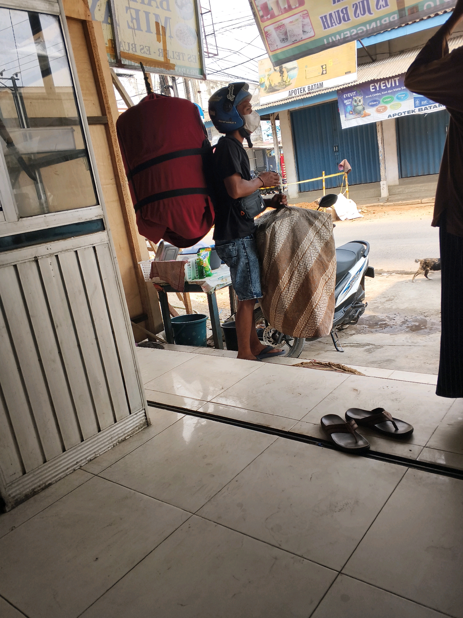 In Sukoharjo III, Indonesia, a man wearing a black t-shirt, denim shorts, a blue helmet, and a face mask stands next to a parked white motorcycle. He is holding a large, red cylindrical object wrapped in a patterned cloth. The motorcycle is covered with a brown and white patterned cloth. In the background, there are buildings with blue roller shutters, and advertisements are visible. To the left of the man, a shop's wooden frame and a closed metal door are present. On the tiled floor in the foreground, a pair of brown flip-flops are placed. A dog is seen walking in the middle ground.