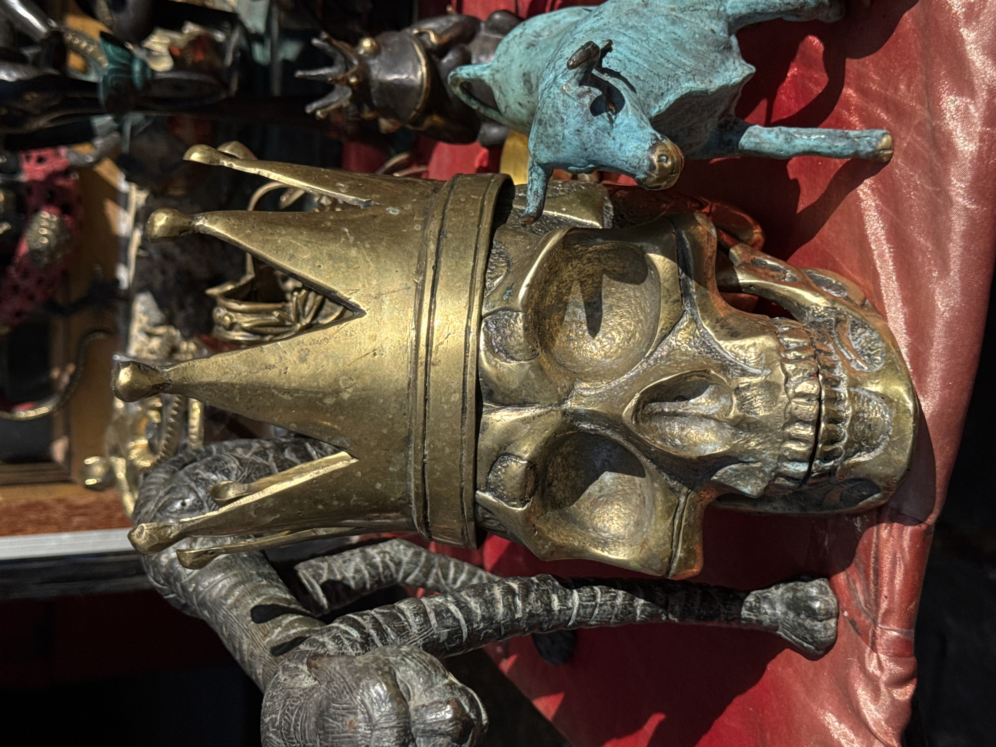 This image shows a collection of bronze statues displayed on a red cloth surface. In the foreground, a large, ornate gold-colored bronze skull wearing a crown is prominently featured. To the left of the skull is a dark bronze statue of a creature, possibly a tiger or a large cat, with intricate detailing on its body and legs. To the right of the skull, a small, turquoise-colored bronze bull stands with its head turned slightly towards the viewer. In the background, other statues are visible, including what appears to be a frog statue with a crown and other dark, unidentifiable figures. The lighting suggests an outdoor or well-lit indoor setting, casting shadows from the statues onto the red cloth. The overall impression is of an art or curio shop display, possibly in a marketplace. The image is taken from a slightly elevated angle, focusing on the central skull and the surrounding sculptures. It is plausible that this scene is located in Ubud District, Indonesia, given the prevalence of such artisanal crafts in that region.