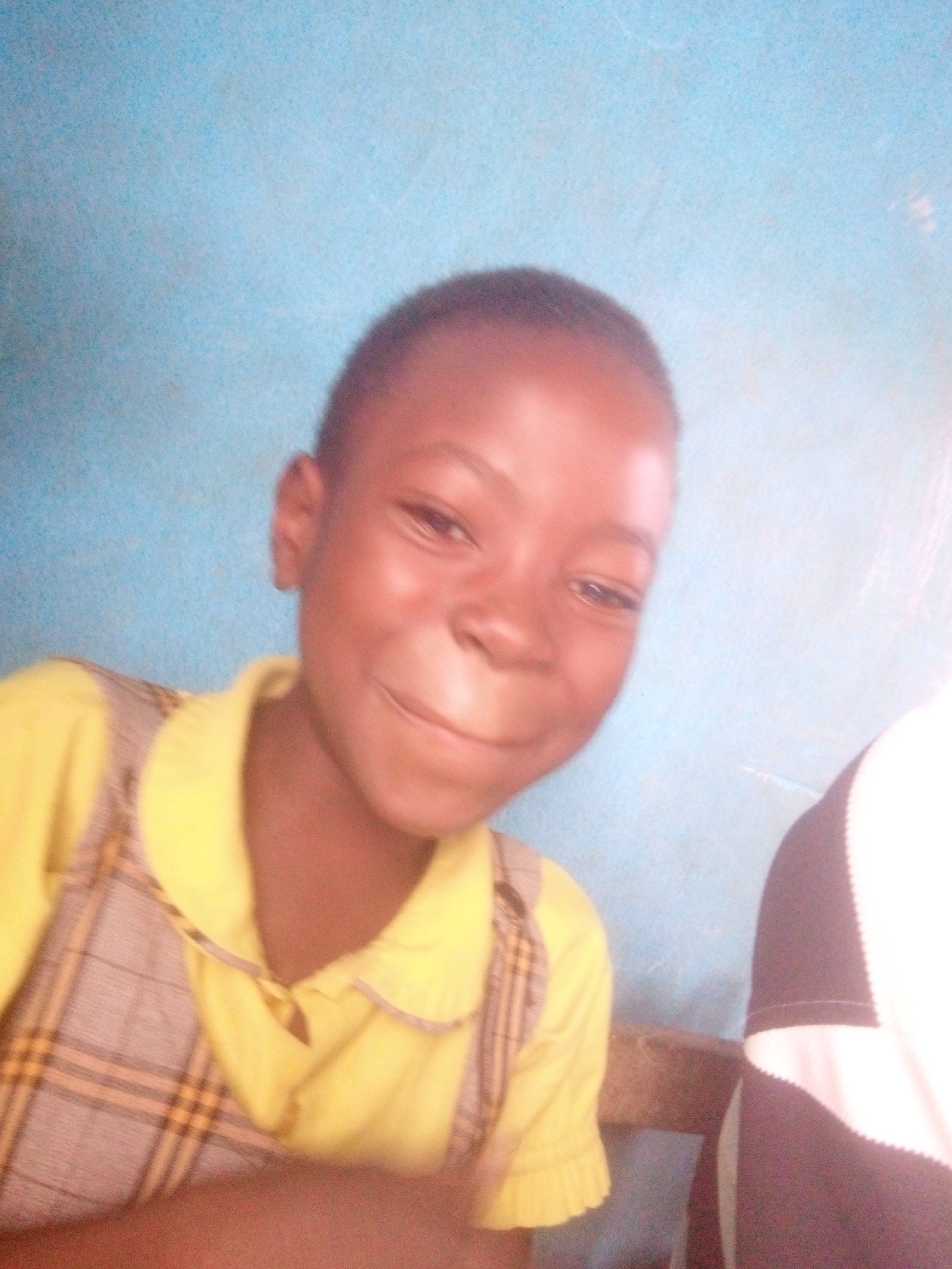 A young Black child with short hair smiles slightly. They are wearing a yellow collared shirt with a grey and yellow plaid pattern on the bib and straps. A small portion of a person wearing a dark and white shirt is visible on the right. The background is a light blue, seemingly a wall. The location is presumably Ikorodu, Nigeria, though this is not evident from the image itself.