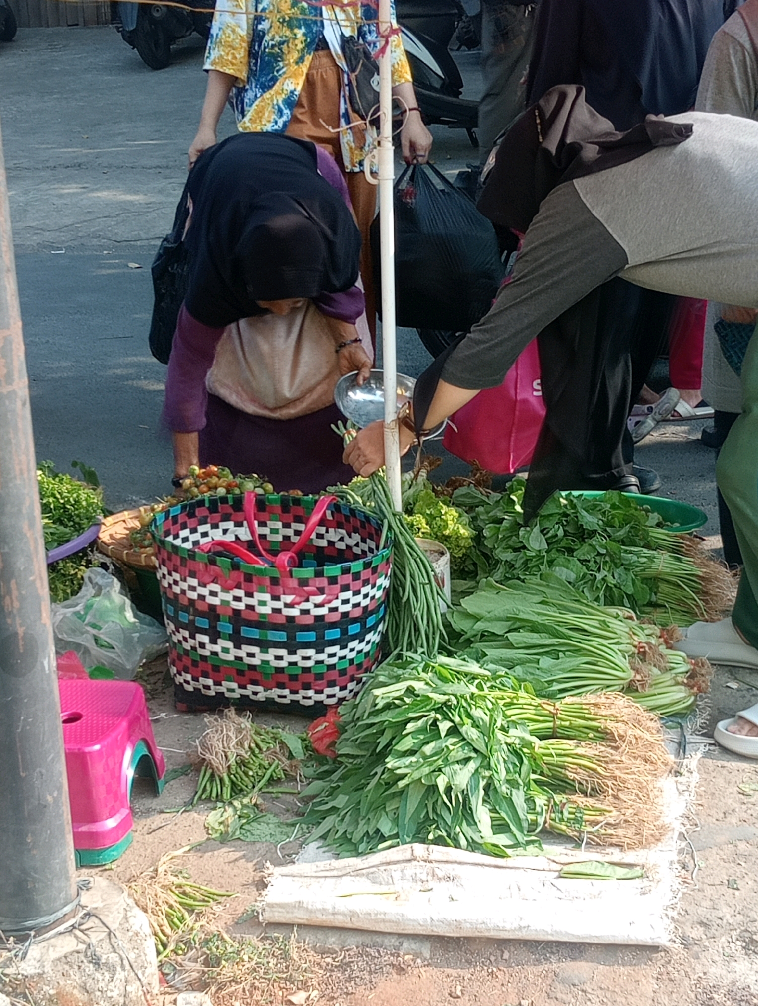 At a roadside market stall in Bandar Lampung, Indonesia, several individuals are engaged in transactions involving fresh produce. A woman wearing a black hijab and a purple tunic is bent over, examining produce in a basket. To her right, a person in a gray long-sleeved shirt and black pants is also reaching for the vegetables. In the foreground, an assortment of vegetables, including long beans and leafy greens, are arranged for sale. A colorful woven basket with pink handles sits prominently. In the background, a motorcycle is parked, and other market-goers can be seen. The scene is set outdoors on a paved surface, indicative of a bustling urban market.