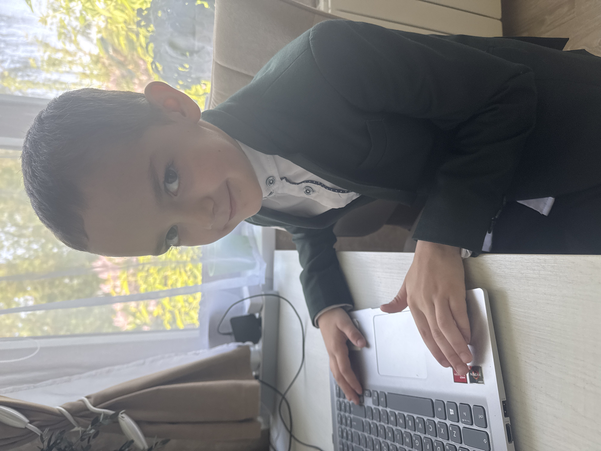 A young boy wearing a dark blazer and a white shirt is seated at a table and using a silver laptop. He is looking towards the camera. A window with white blinds and a tree outside is behind him. The location is Раздолье, Russia.