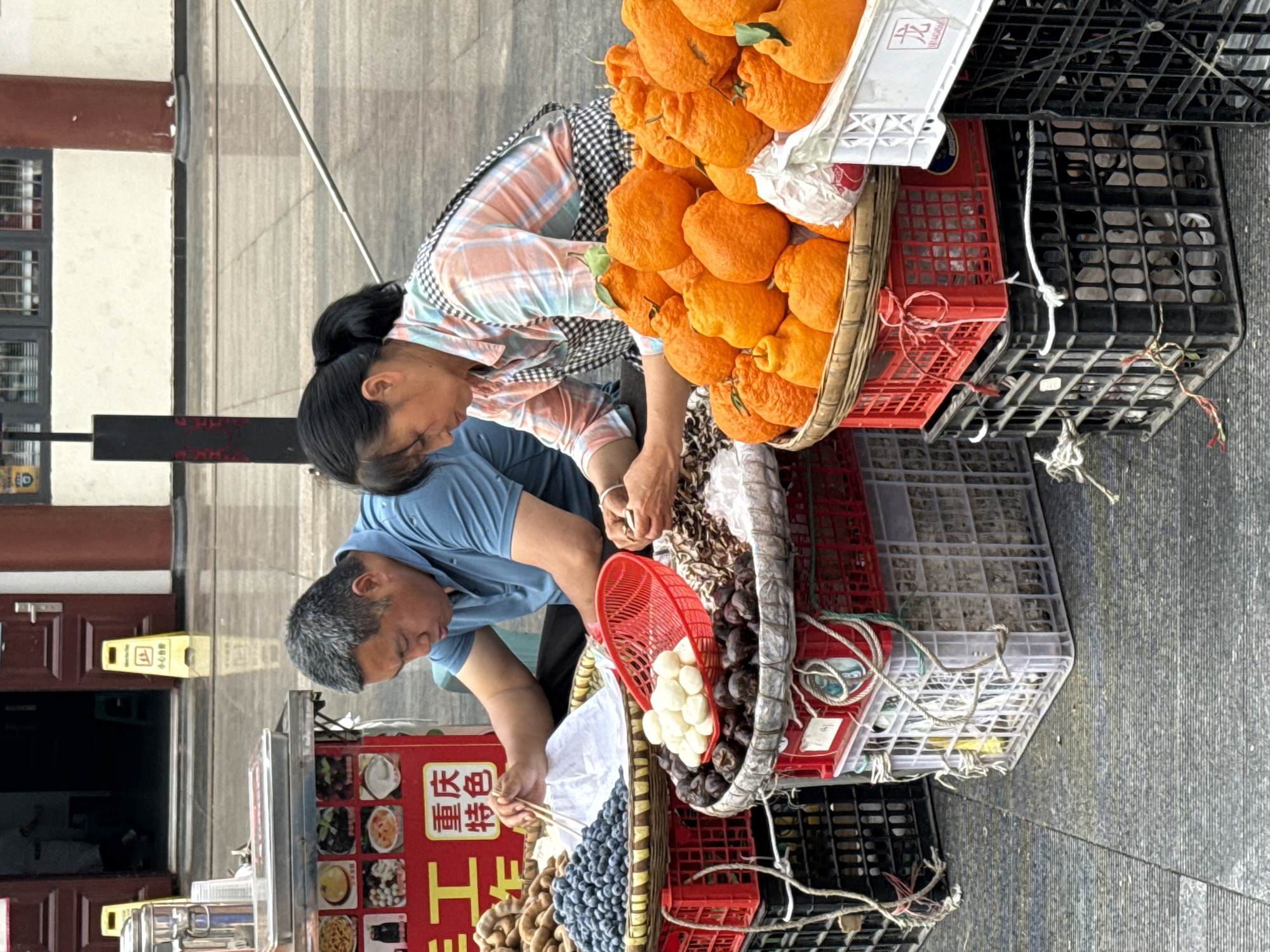 Two adults are crouched over a fruit and vegetable stand on a city street in Yuzhong District, China. The stand consists of baskets and plastic crates stacked on top of each other, displaying tangerines, chestnuts, water chestnuts, and blueberries. One adult is wearing a light blue shirt and is using chopsticks to pick up some food. The other adult is wearing a plaid shirt. The background includes a building with a yellow "wet floor" sign near the entrance, and a menu board with food items.