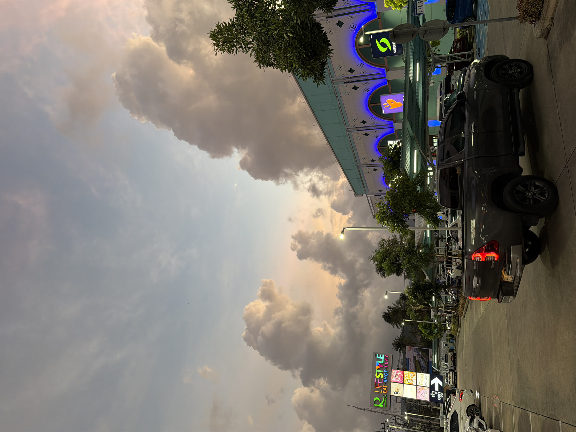 In Si Sunthon, Thailand, a grey pickup truck is parked in a lot in front of a building. The sky is filled with large, dramatic clouds that are illuminated by the setting sun, casting a warm glow. The building is modern, with a light blue facade and arched windows that are lit with blue neon lights. A large sign with colorful text that reads "R LIFESTYLE ROBINSON EAT. SHOP. PLAY." is visible on the left side of the image. Several other cars are parked in the lot, and trees and streetlights are also present. The scene appears to be a commercial area at dusk.