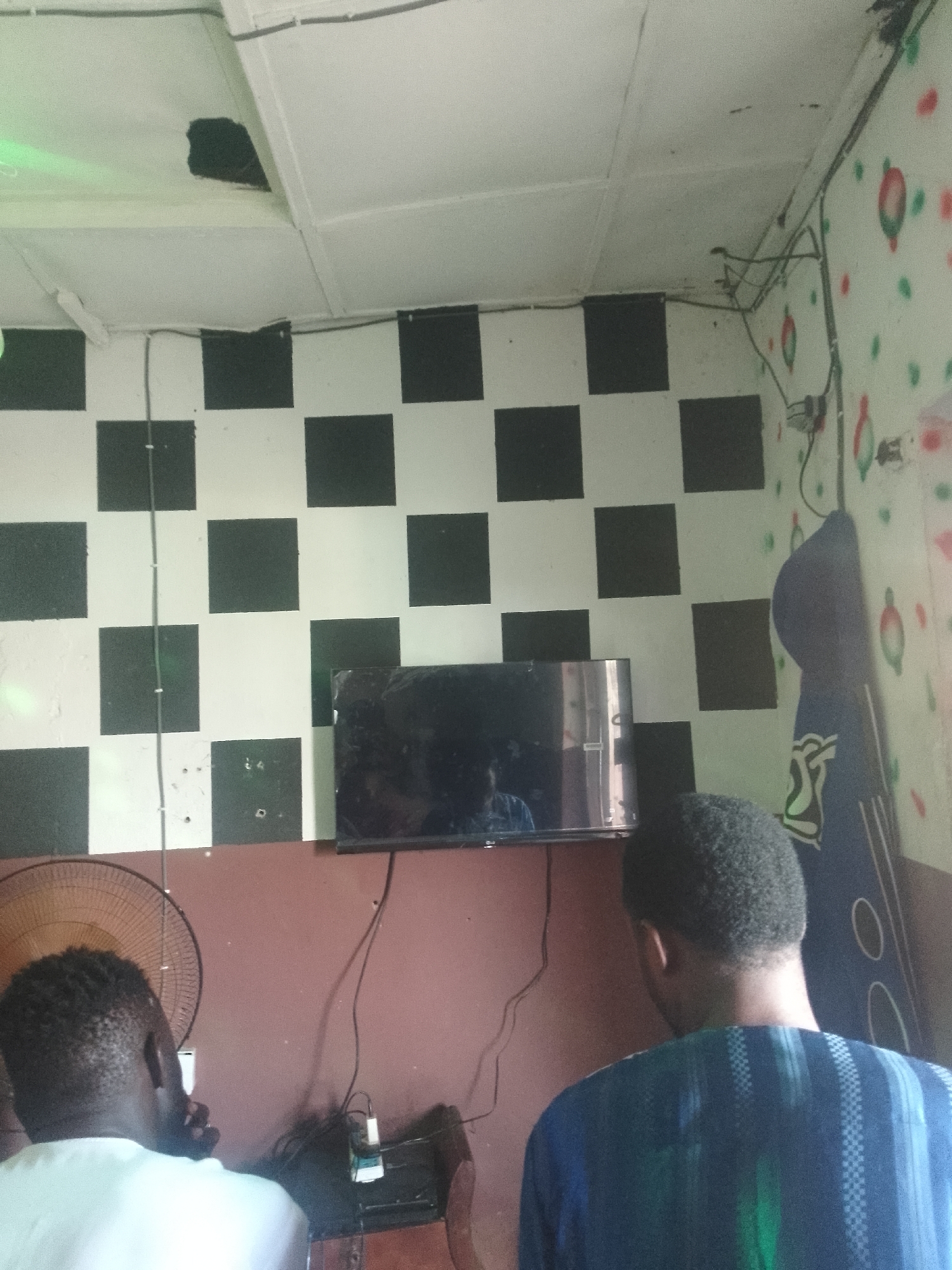 The image depicts an indoor scene, likely a room, in Serti, Nigeria. The walls are adorned with a checkerboard pattern of black and white squares, and another section of the wall has a pattern of red and green circular shapes. A television is mounted on the wall, and two individuals, both appearing to be male with dark skin and short, dark hair, are positioned in front of it. One individual, seen from the back and shoulder, is wearing a white shirt. The other individual, also seen from the back, is wearing a blue garment with a striped pattern. In the foreground, a large, round fan with a brown grille is visible on the left side of the image. Electrical wires are present, running along the walls and ceiling. There appears to be a hole or damaged section in the ceiling, revealing darkness within. A dark-colored object, possibly a guitar case or a bag, leans against the wall on the right. The overall environment suggests a casual or recreational space.