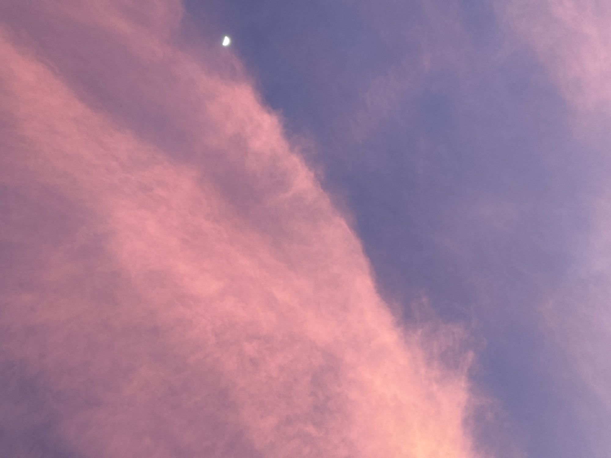 The image shows a sky at twilight or dusk in Mũi Né, Vietnam. A crescent moon is visible in the upper-center of the frame. The sky is divided into two main color segments: a pinkish, cloud-filled area on the left and a darker, purplish-blue area on the right. The clouds are wispy and arranged in diagonal patterns. The overall lighting suggests a soft, diffused light source, likely the setting or rising sun. No people or distinct objects are visible other than the sky and the moon.