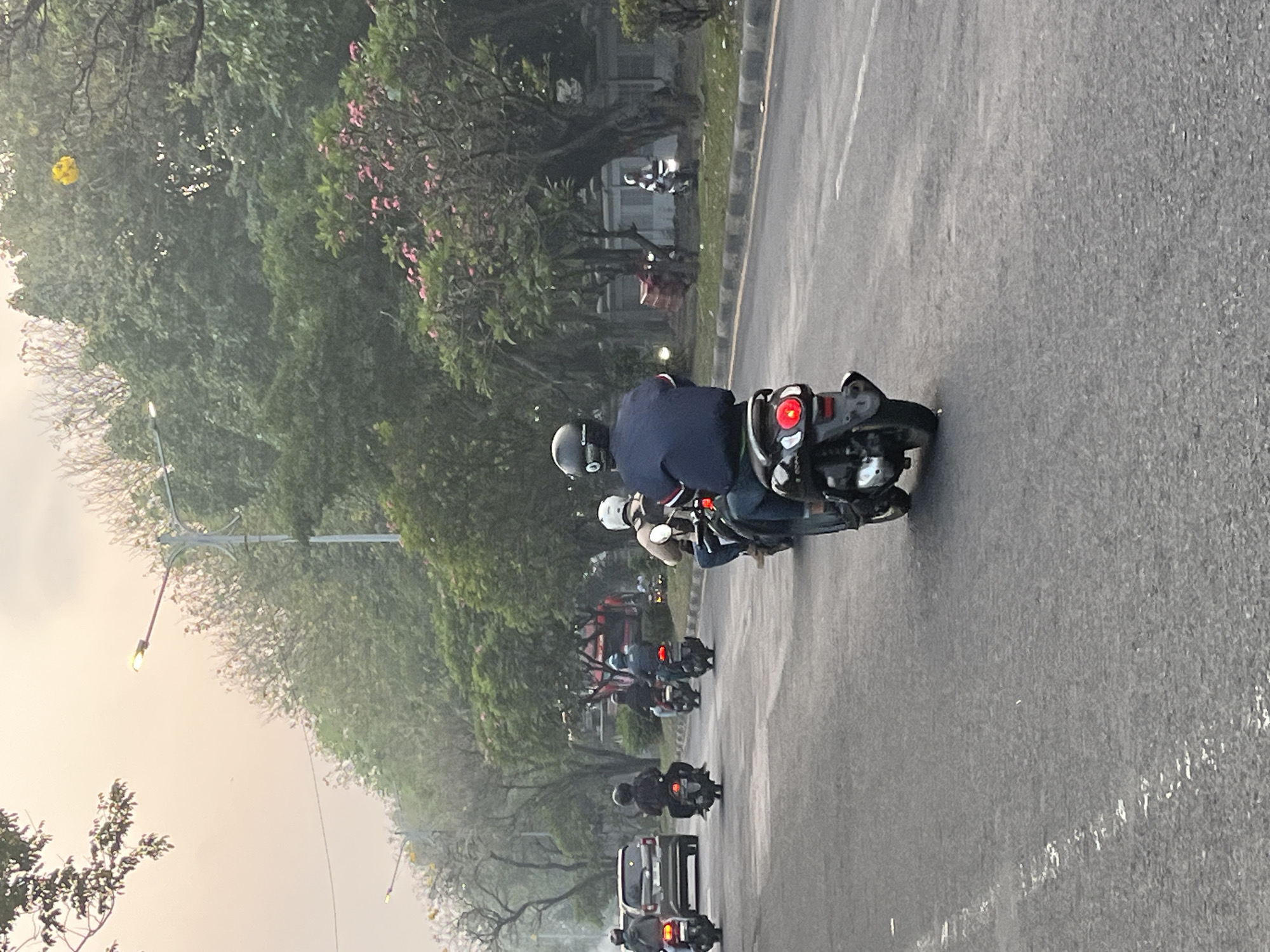 In Sumorame, Indonesia, an overcast sky casts a diffused light over a busy road lined with lush green trees. Several motorcycles, each with one or two riders wearing helmets, traverse the asphalt. One motorcycle, in the foreground, has a rider in a dark blue jacket and jeans, his helmet is black with silver accents. Behind him, another motorcycle with two riders is visible. Further down the road, a silver car drives in the same direction as the motorcycles. The environment is characterized by dense foliage and trees, with some displaying pink blossoms. Streetlights are present along the roadside, their yellow lights faintly illuminating the scene. A yellow object, possibly a balloon, floats amongst the tree branches in the distance. The road is marked with white lines, indicating lanes of traffic.
