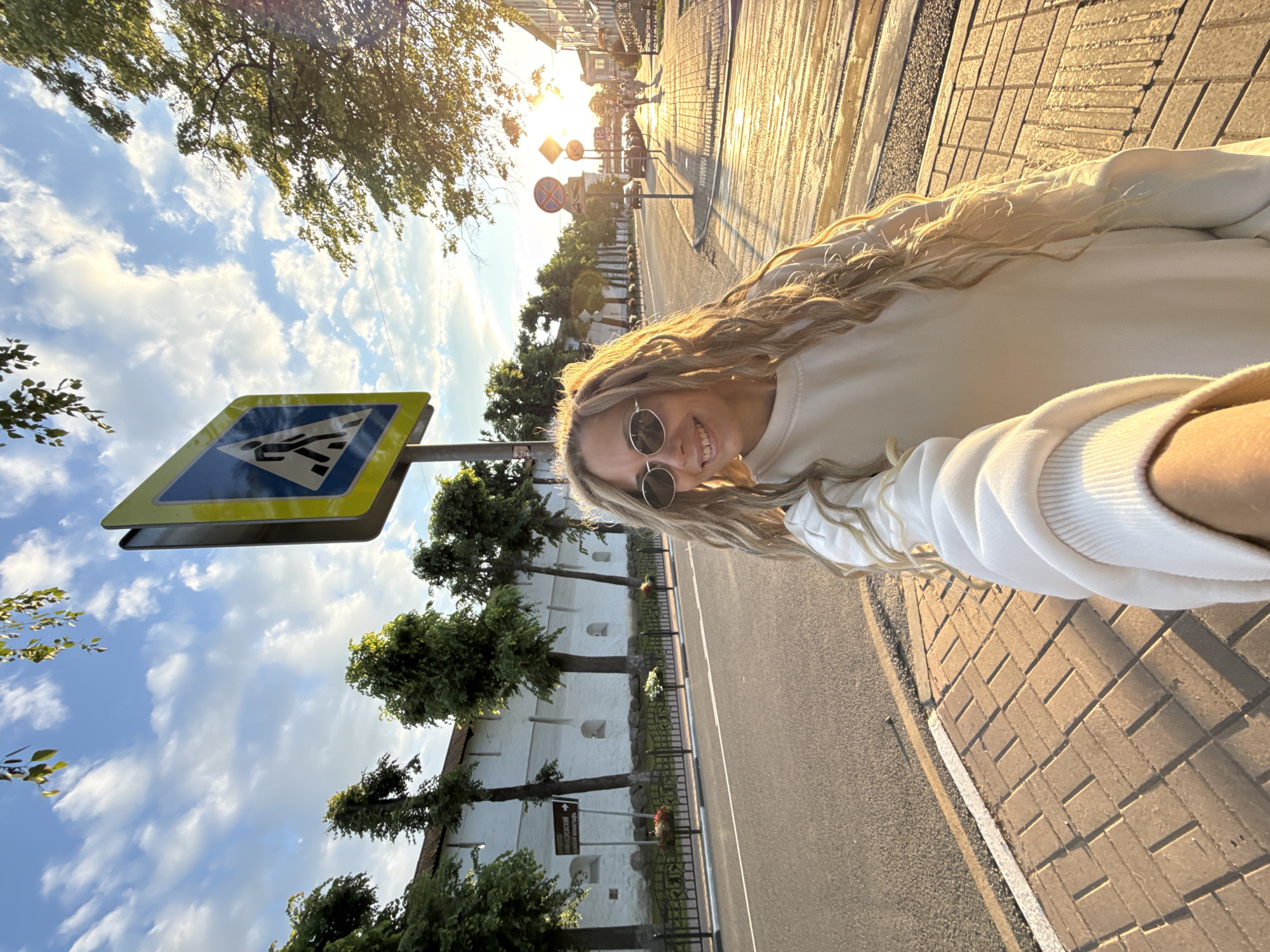 A young woman with long, wavy blonde hair and round sunglasses is taking a selfie outdoors, smiling at the camera. She is wearing a light-colored sweatshirt. The image is taken from a low angle, showing a bright, sunny scene during what appears to be golden hour, with warm light and long shadows.
In the foreground, a prominent blue and yellow pedestrian crossing sign is visible to the left of the woman. Behind her, a paved sidewalk with brick-like patterns extends alongside an asphalt road. Across the road, there is a distinctive long, white wall featuring several arch-shaped niches. This wall is lined with trees and a black metal fence. A brown directional sign near the wall, partially visible, indicates "БЛАГОВЕЩЕНСКИЙ" (Annunciation), likely part of a street or area name. In the distance, other buildings and street signs, including a prohibitory sign with a red circle and diagonal line, can be seen along the street. The sky above is blue with scattered white clouds. The overall setting is an urban or historical environment, consistent with Заволжье, Russia. The woman appears happy and relaxed.