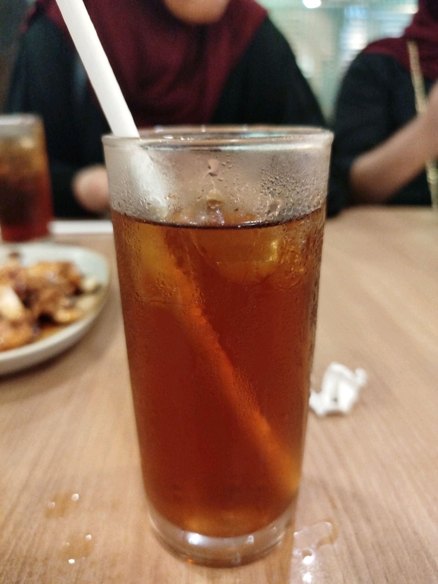 A close-up, eye-level shot features a glass of iced tea on a wooden table. The glass is condensation-covered and contains a white straw and ice cubes. In the background, out of focus, two individuals wearing dark clothing and maroon headscarves are visible, suggesting a restaurant or cafe setting in Balikpapan, Indonesia. A plate with food items is also partially visible to the left of the glass.