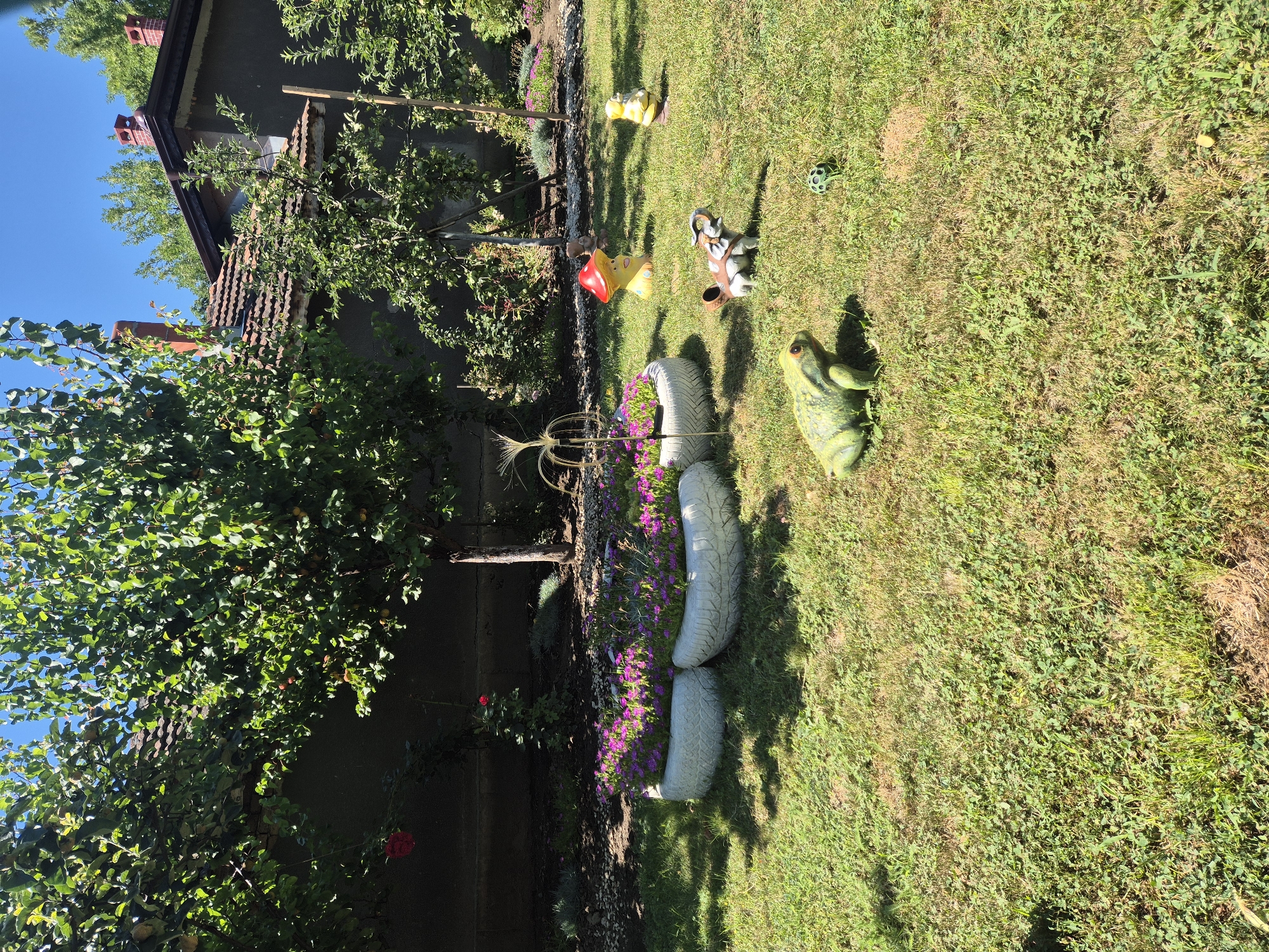 An outdoor, eye-level, full shot shows a garden with a residential building in the background on a sunny day. In the foreground, a large, green, ceramic frog is positioned on a grassy lawn. To the right of the frog, a small, green, patterned ball rests on the grass. Further back, a collection of garden gnomes is scattered across the lawn. These include a mushroom-shaped gnome with a red cap and a smiling face, a yellow chick-like gnome, and a grey ceramic elephant with a rider. In the mid-ground, two large, white tire planters filled with vibrant purple flowers are situated. Behind the planters, a large tree with green leaves dominates the scene, its branches extending towards the top of the frame. Behind the tree, a dark, textured wall is visible. The residential building in the background has a tiled roof and chimneys. The sky above is a clear, bright blue.