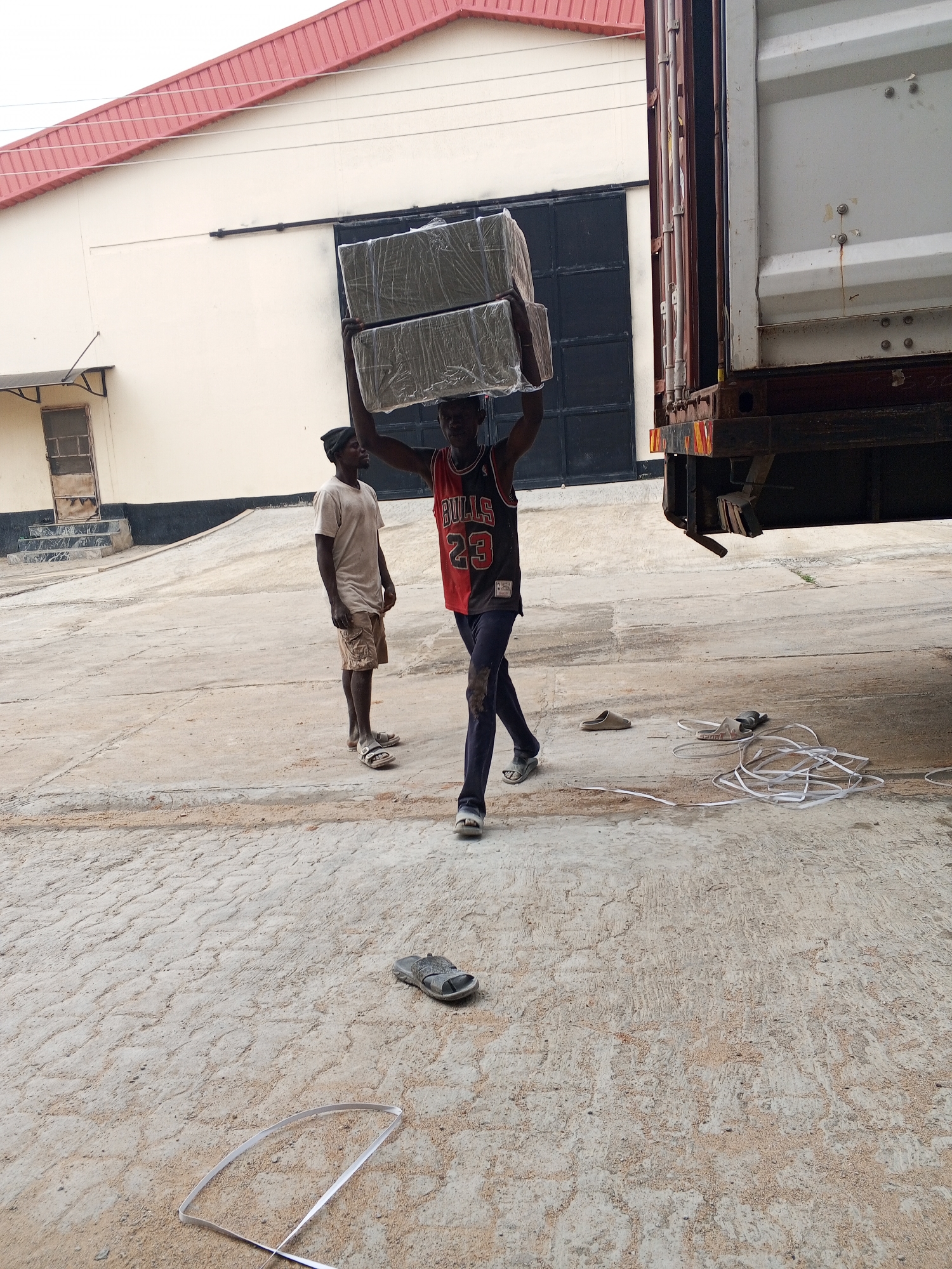 In Pasere, Nigeria, a man in a "Bulls 23" jersey carries two large, wrapped rectangular objects on his head, heading towards a shipping container attached to a truck. Another man, wearing a t-shirt and shorts, stands nearby watching. A pair of sandals lies discarded on the concrete ground, along with a pile of white strapping material and a long piece of strapping. In the background, there is a beige building with a red roof and a large metal door.
