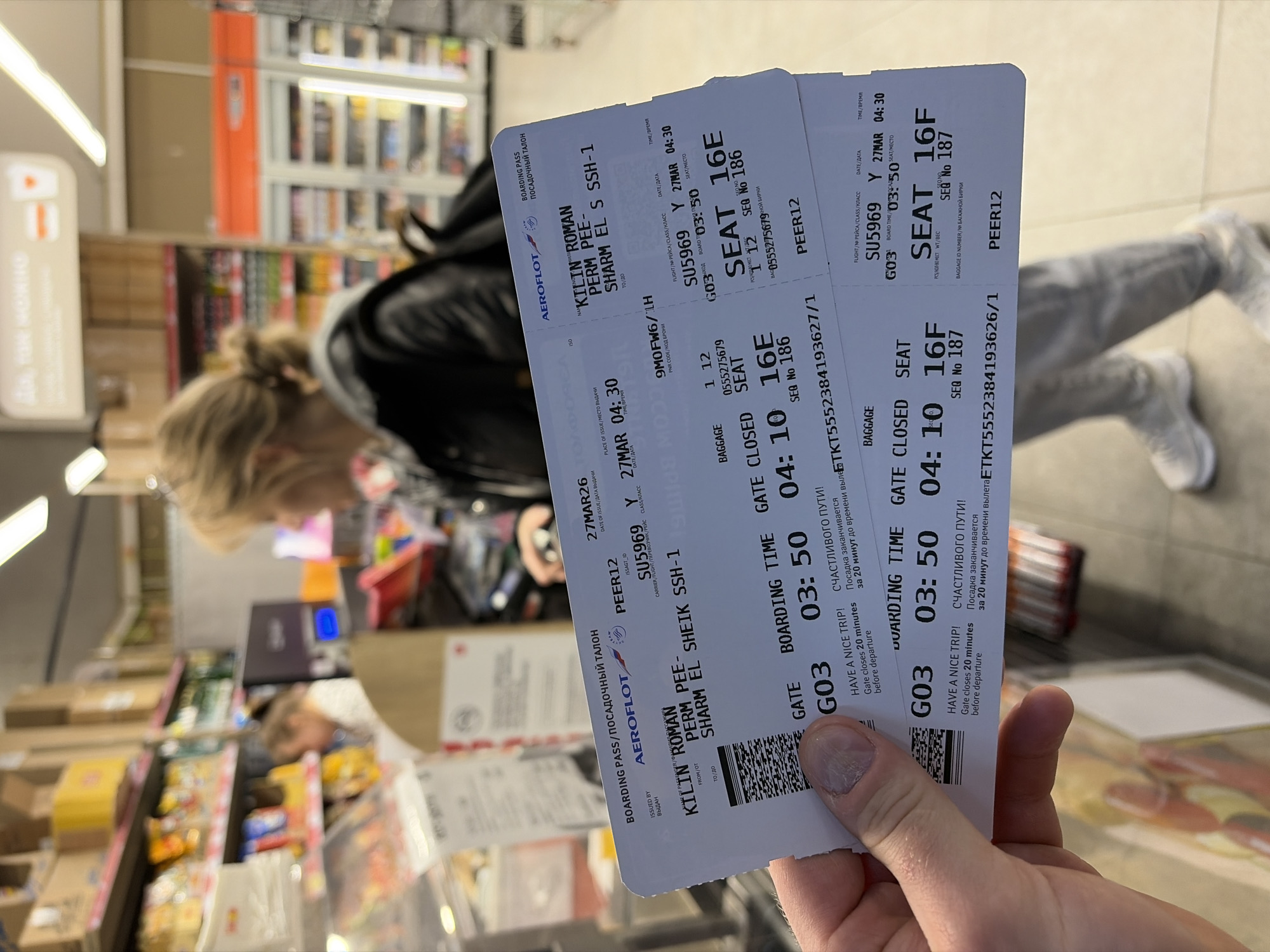 The image depicts an indoor retail environment, consistent with a convenience store or mini-market within an airport terminal. The location is not in Boldino, Russia, as the visible boarding passes indicate a flight route from Perm, Russia (PEE) to Sharm El Sheikh, Egypt (SSH-1).
In the foreground, a light-skinned left hand holds two white Aeroflot boarding passes. The thumb, index, and middle fingers are visible, with short, unpolished nails. The boarding passes are for passenger KILIN ROMAN on flight SU5969, departing on March 27th, with a boarding time of 03:50 at Gate G03, for seats 16E and 16F. The destination is Sharm El Sheikh, originating from Perm. Each pass displays a barcode, "HAVE A NICE TRIP!" in English, and "СЧАСТЛИВОГО ПУТИ!" in Russian.
In the mid-ground, a woman with blonde hair tied back in a bun is seen from behind, facing away from the camera and slightly to the right. She wears a dark (likely black) jacket with a grey hood visible at the neckline, light-colored pants, and white sneakers. She appears to be standing or moving in an aisle.
The background features extensive retail shelving units stocked with various consumer goods. On the left, shelves hold packaged snacks (e.g., chips, candies) and other items, with some products in cardboard boxes stacked above. Further back on the left, a blurry figure with light hair, wearing a light-colored top, is partially visible behind a counter or service area, possibly a checkout. On the far right, a tall, clear-doored commercial refrigerator is visible, containing bottled items. Overhead fluorescent or LED strip lighting illuminates the entire scene. The floor is light-colored.