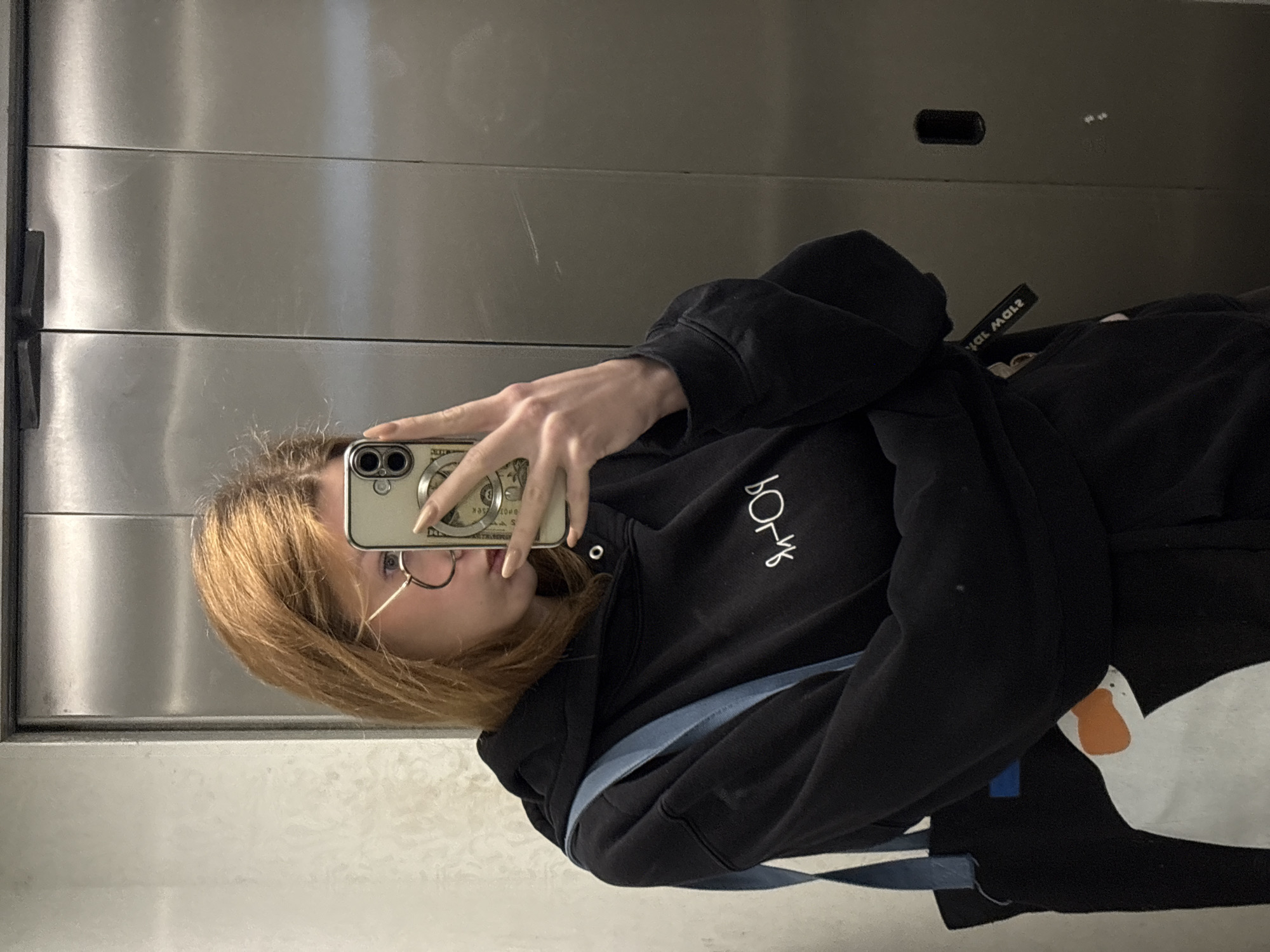 A young person with light brown hair and glasses is taking a selfie in what appears to be an elevator. They are wearing a black hoodie with white lettering on the chest and a blue bag strap is visible across their body. The phone case has an image of money on it. The elevator walls are brushed metal. The image does not contain information that would confirm a location in Kragujevac, Serbia.