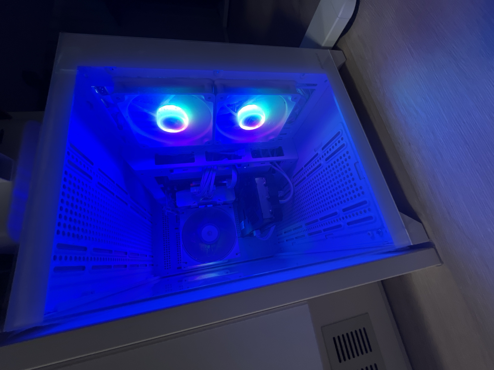 The image shows an open computer case with internal components illuminated by blue RGB lighting. The computer case is white and is positioned on a wooden floor. Two RGB fans are visible on the front of the case, displaying a gradient of colors from purple to green and blue. A larger fan is visible on the back of the case, also illuminated. A black heatsink with attached fan is visible in the middle of the case. The computer case is standing upright. The background is dark and out of focus. The scene is indoors. There are no people present in the image. The location of Varna, Bulgaria is not determinable from the image content.