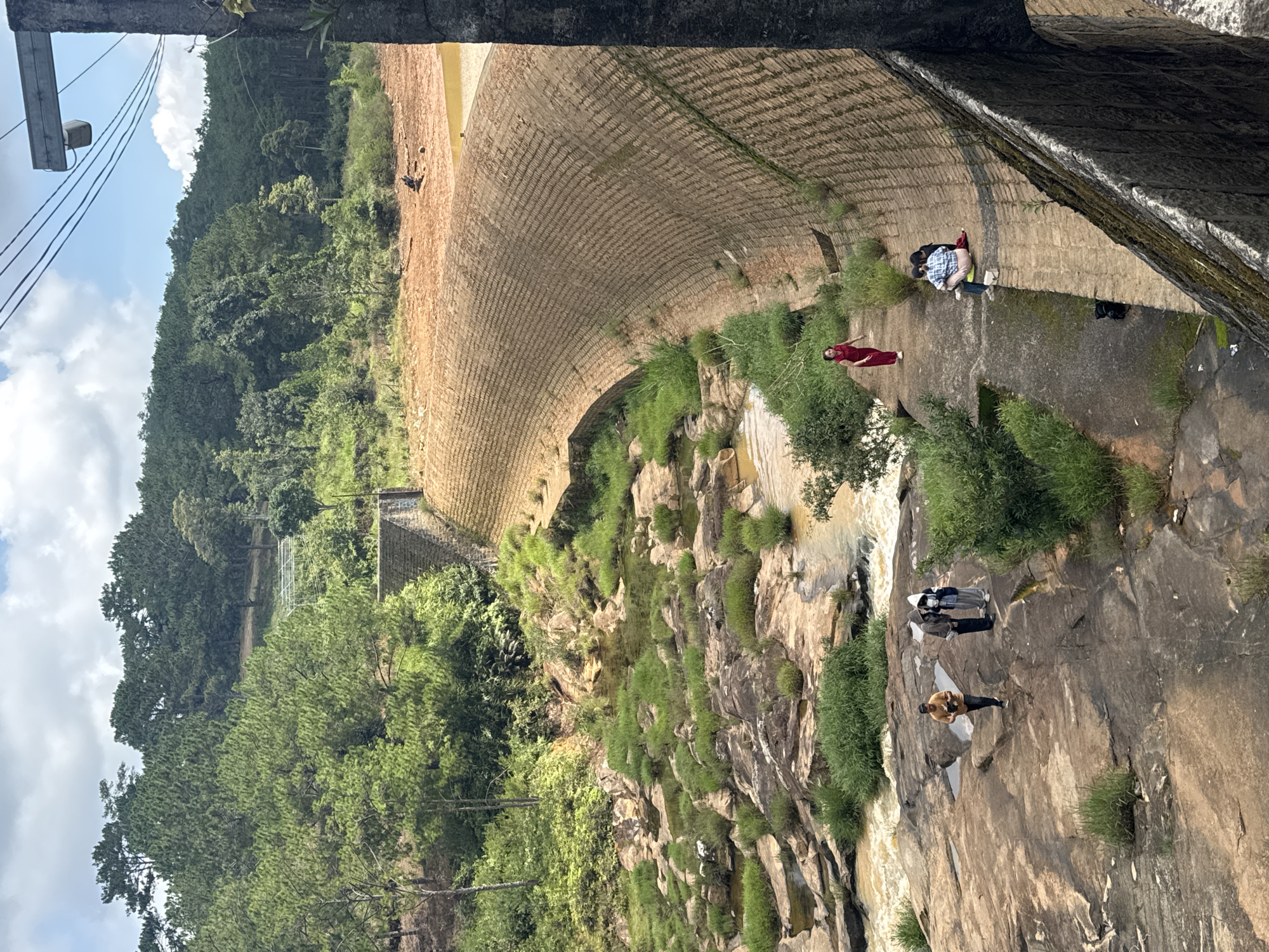 A curved concrete dam with a rough, textured surface is the dominant feature in the foreground and midground of the image, set against a backdrop of lush green hills and trees. The dam's surface is patterned with numerous small, rectangular indentations, forming a repeating grid-like design. On the flat area near the base of the dam, several people are visible. One woman in a red dress walks along a path to the left. Further to the right, a group of three people are seated on the dam's surface, one wearing a plaid shirt. On a rocky outcrop to the left of the dam, a group of three individuals stand, one holding a camera. The water below the dam flows over rocks, creating a stream with surrounding greenery. The background consists of a dense forest of pine trees covering rolling hills. Power lines stretch across the top right of the frame, connected to a concrete structure. The sky is partly cloudy with patches of blue. The scene appears to be an outdoor recreational area, possibly a tourist attraction, located in Lang Biang - Da Lat, Vietnam, given the distinctive dam architecture and surrounding natural landscape.