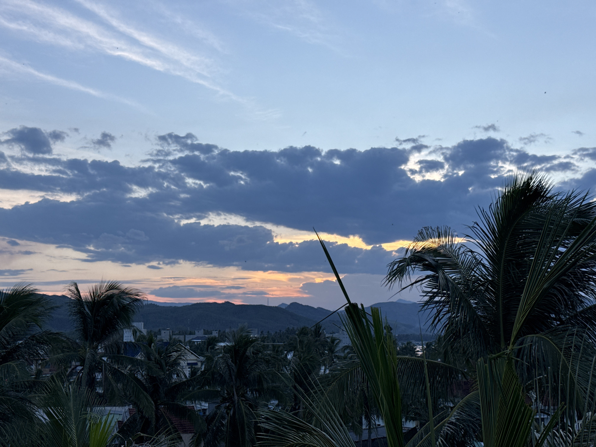 The image depicts a twilight scene, possibly from a balcony or elevated viewpoint, overlooking a residential area with lush palm trees in the foreground. The sky is a gradient of blues, with streaks of white cirrus clouds and darker, more substantial clouds lower in the frame. The setting sun casts a warm, orange glow behind a range of distant, dark mountains. Silhouettes of palm fronds dominate the foreground, obscuring parts of the town below, which consists of buildings with visible roofs and structures. The overall mood is serene and tranquil, characteristic of dusk.
