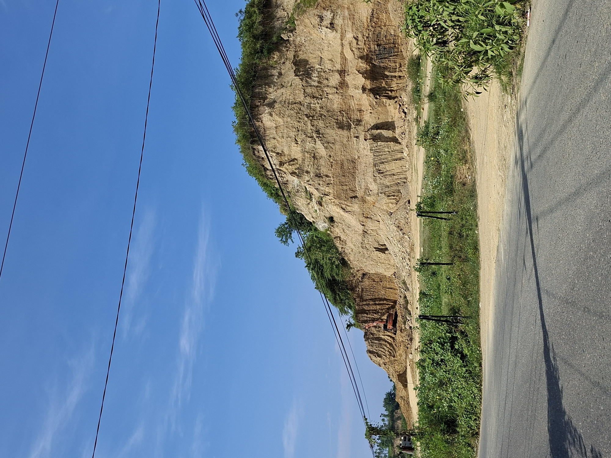 The image, taken in Lhokseumawe, Indonesia, captures a bright, sunny outdoor scene featuring an excavated landscape alongside a road. On the right side, an asphalt road runs parallel to a lighter dirt track or path. To the left of this path, a massive, towering dirt and rock cliff dominates the midground, showcasing distinct horizontal layers and textures indicative of extensive excavation or natural erosion.
Green vegetation, including bushes and small trees, grows on the top edge and at the base of the cliff, as well as intermittently along the dirt path and beside the paved road. In the mid-distance, nestled at the base of the cliff, a piece of heavy machinery, likely an excavator, with a reddish-orange color, is visible. Overhead, several dark electrical or telephone wires stretch across the clear, vibrant blue sky, which contains only a few wispy white clouds.
Two slender dark poles stand near the edge of the dirt path and road. Strong shadows are cast upon the paved road from an unseen object to the right, suggesting it is midday or early afternoon. The scene is devoid of people. There is no visible text. The overall impression is one of active land transformation or a natural landscape modified by human intervention, under pleasant weather conditions.