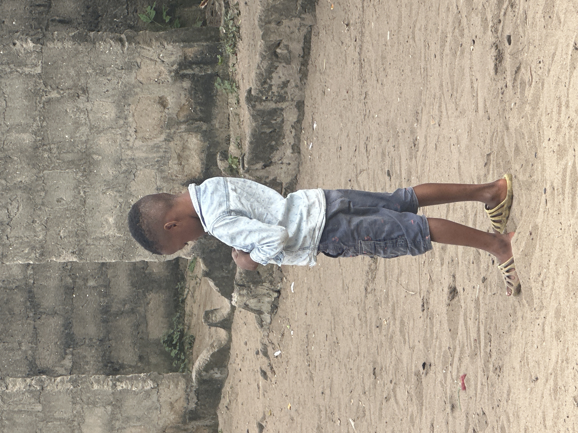 A young boy, viewed from the side, stands on sandy ground in front of a rough concrete wall in Badagry, Nigeria. He is wearing a light blue denim shirt and dark grey cargo shorts. His head is bowed as he looks down, his hands clasped together in front of him. He wears light-colored sandals on his feet. The environment appears somewhat unkempt, with patches of greenery growing near the base of the wall and debris scattered across the sandy surface. The concrete wall is textured and weathered, suggesting age and exposure to the elements. The lighting is overcast, casting a soft, diffused light on the scene.