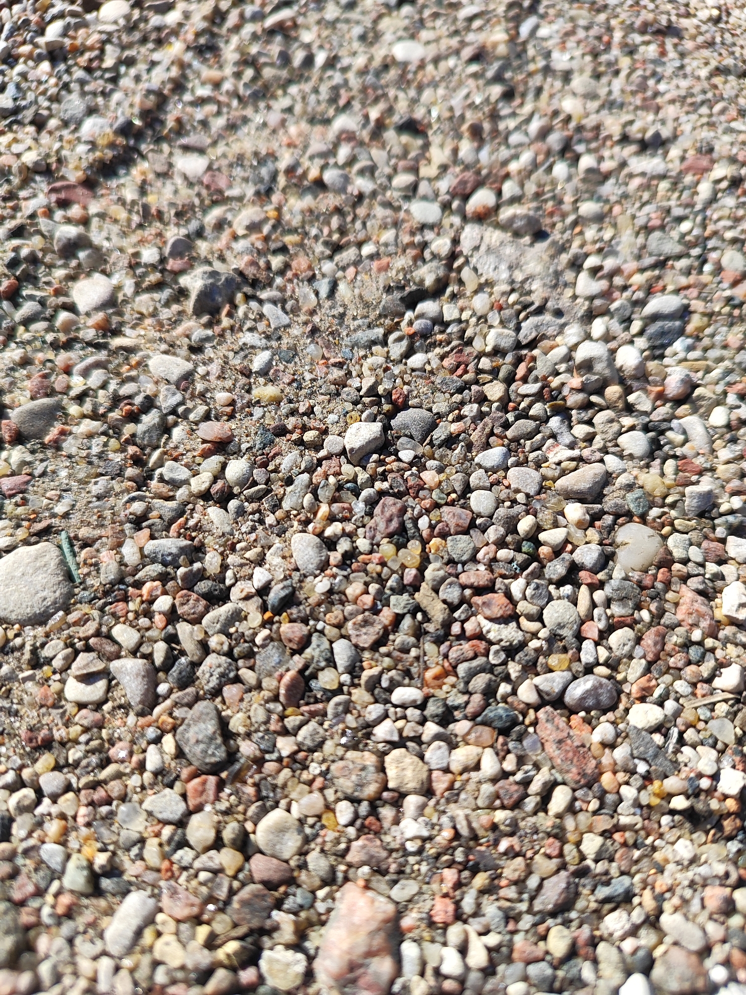 The image shows a close-up, overhead view of a gravel surface. The gravel consists of small, multi-colored stones and sand. The stones vary in size, shape, and color, with shades of gray, brown, red, and white visible. Some stones appear smooth and rounded, while others are more angular. The lighting suggests it is daytime, with shadows cast by some of the larger stones. The ground is dry. There are no people or animals visible in the image. The image is likely a photograph of a section of a road, path, or outdoor area covered in gravel. Without further information, it is not possible to determine the specific location within the city of Hrodna, Belarus.