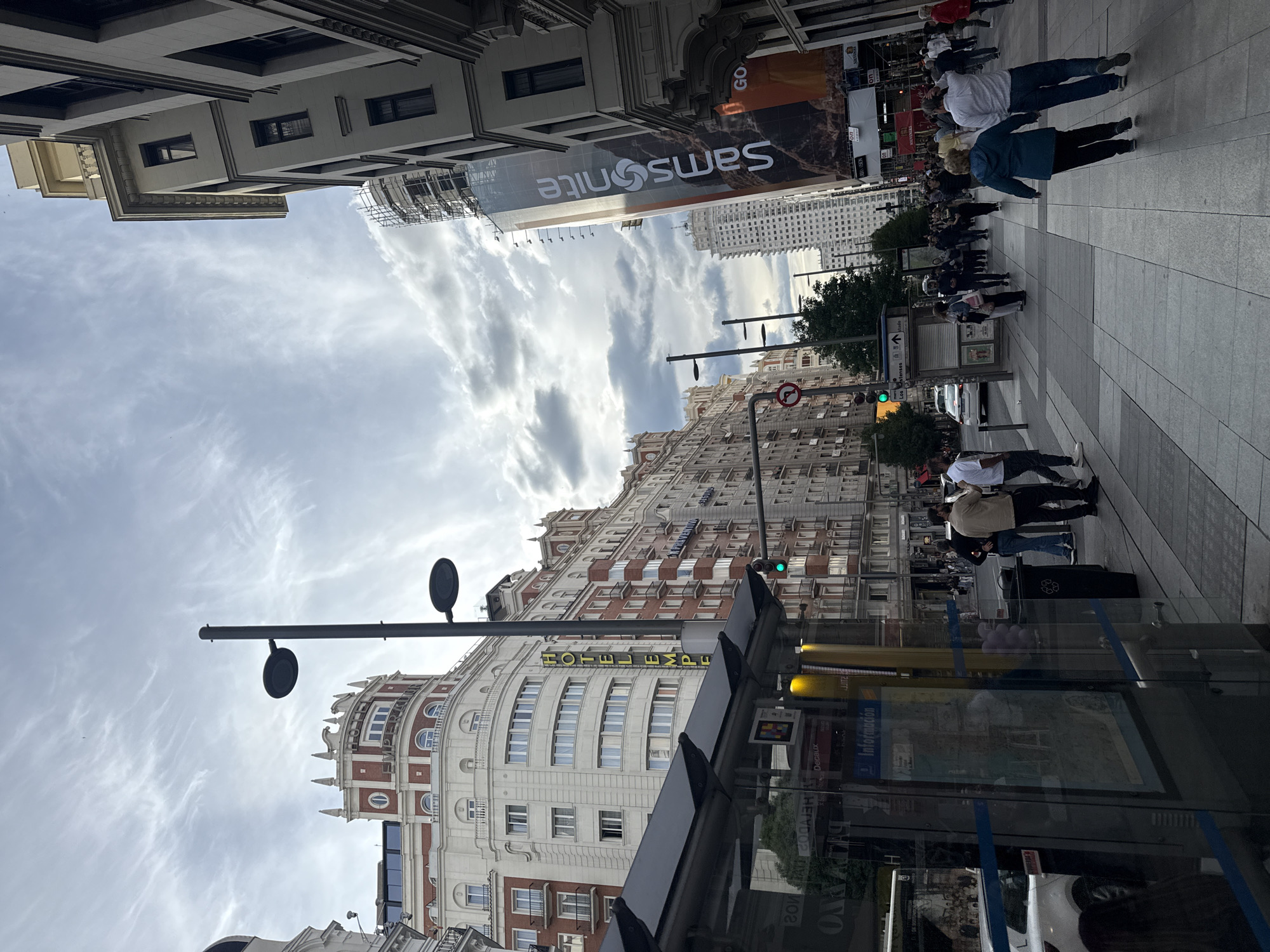 The image shows a street view in Madrid, Spain. The focus is on the architecture, with several buildings visible, including the Hotel Emperador, identifiable by its yellow signage. Above, a large "Samsonite" advertisement is displayed on a building. Pedestrians are present on the sidewalk, and traffic lights are also visible. Street furniture such as lamps is in view. The sky is cloudy, providing an overcast ambiance. The presence of a bus stop with information displays suggests a public transport route. The angle of the shot gives a unique perspective of the urban landscape.