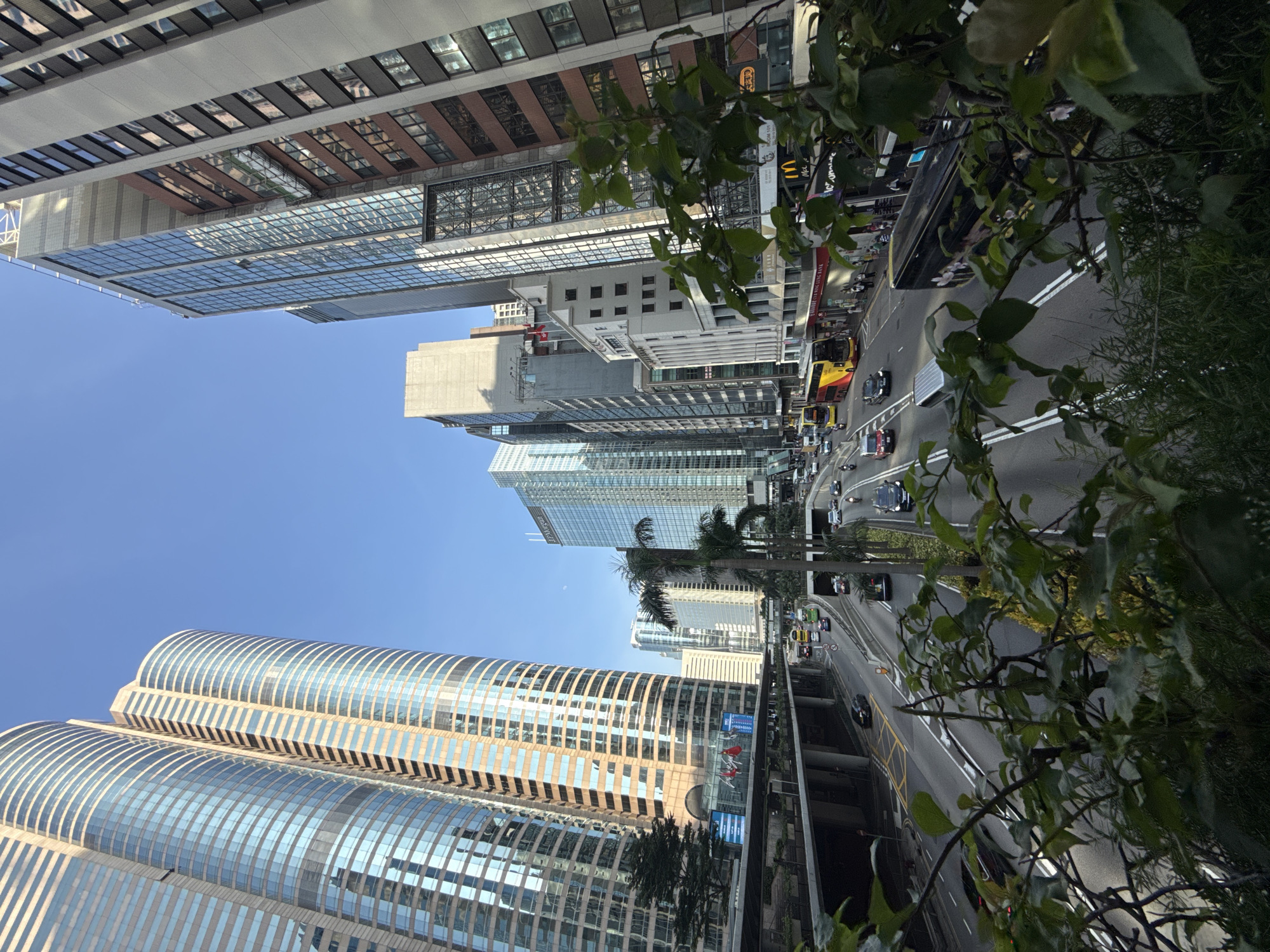 The image captures a bustling street scene in Hong Kong, China, dominated by towering modern skyscrapers on a clear, sunny day. The vantage point is elevated, looking down onto a multi-lane road filled with traffic.
Prominently featured are the architectural marvels of Hong Kong's financial district. On the left, two curved, glass-clad skyscrapers with beige horizontal accents rise majestically against the blue sky. These buildings reflect the surrounding environment and the blue sky. To the right, a variety of other skyscrapers with different architectural styles, including glass, concrete, and brick facades, form a dense urban landscape. One notable building has a prominent "J.P. Morgan" sign.
The road below is a hive of activity. Several double-decker buses, a common sight in Hong Kong, are visible, including one yellow and red bus and a black bus with floral motifs on its side. Cars, including red and black sedans, and a silver van, navigate the lanes. A motorcycle is also present in the traffic. Pedestrians are visible on the sidewalks, and some establishments, including a McDonald's, are marked by their signage.
The environment suggests a well-developed urban infrastructure, with overpasses and elevated walkways visible above the road. Lush green foliage, including trees and leafy branches in the foreground, provides a natural contrast to the urban setting, suggesting the presence of green spaces within the city. The overall impression is one of a vibrant, dynamic, and densely populated metropolitan area.