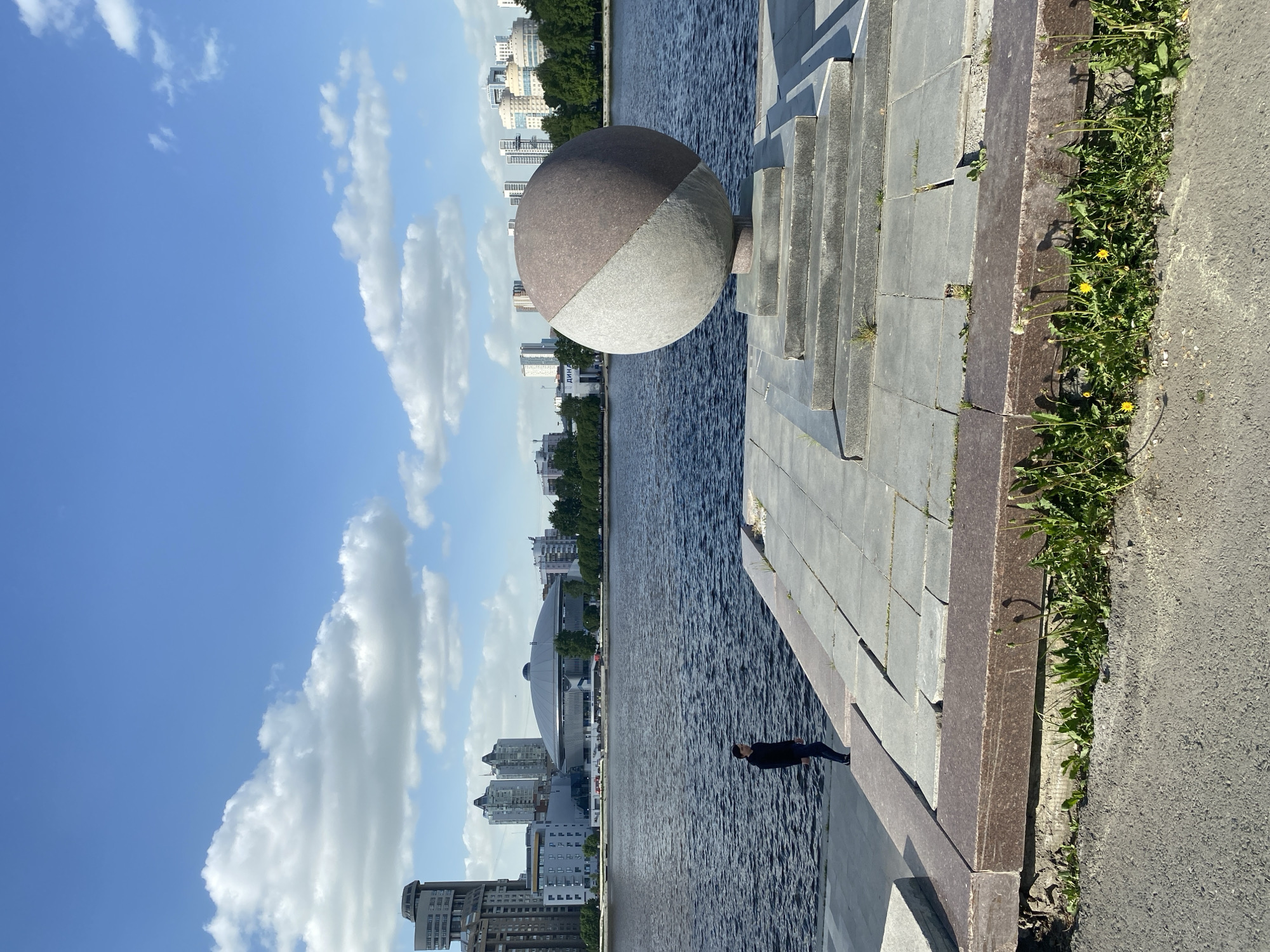 The image shows a waterfront scene in Yekaterinburg, Russia, likely along the Iset River. The sky is blue with scattered clouds. Along the riverbank, there is a stone embankment with grass and weeds growing along the edge. A large, two-toned spherical sculpture sits on the embankment. In the water, a figure dressed in black is lying, possibly floating. In the background, buildings and trees line the river, including the sports arena. The location is likely in central Yekaterinburg.
