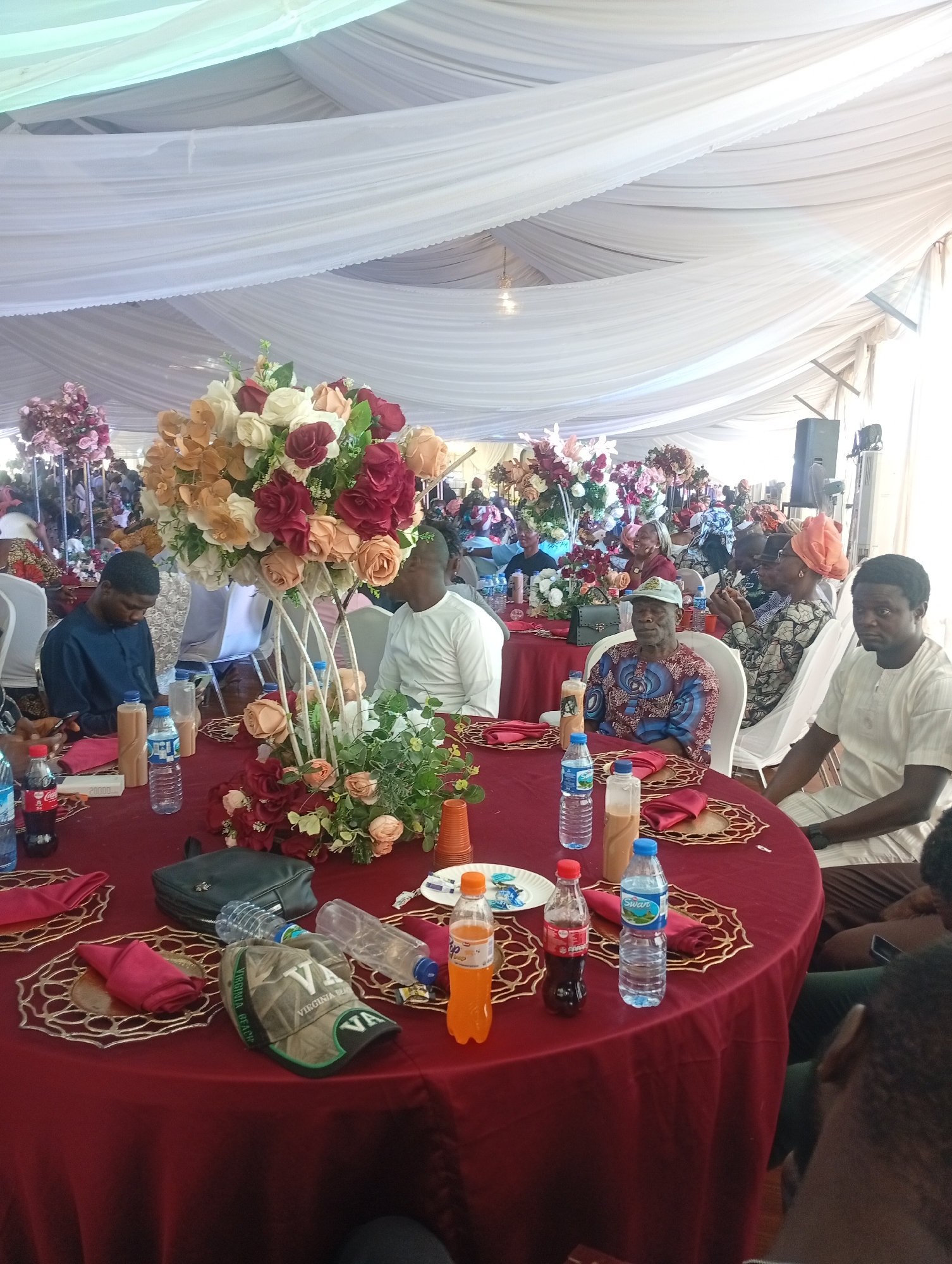 The image shows an indoor event, likely a wedding or celebration, taking place under a large tent draped with white fabric. Multiple round tables covered with red tablecloths are arranged throughout the space, each adorned with centerpieces of flowers in shades of white, red, and orange. Guests are seated around the tables, dressed in a variety of attire ranging from traditional patterned outfits to more modern clothing. Water bottles and various beverages sit on the tables. In the background, additional floral arrangements can be seen, along with speakers suggesting a sound system setup. Given the prompt constraints, I can infer that the location is within the city of Barakin Lerawi, Nigeria, even though the picture has no indicators that confirm the location directly.