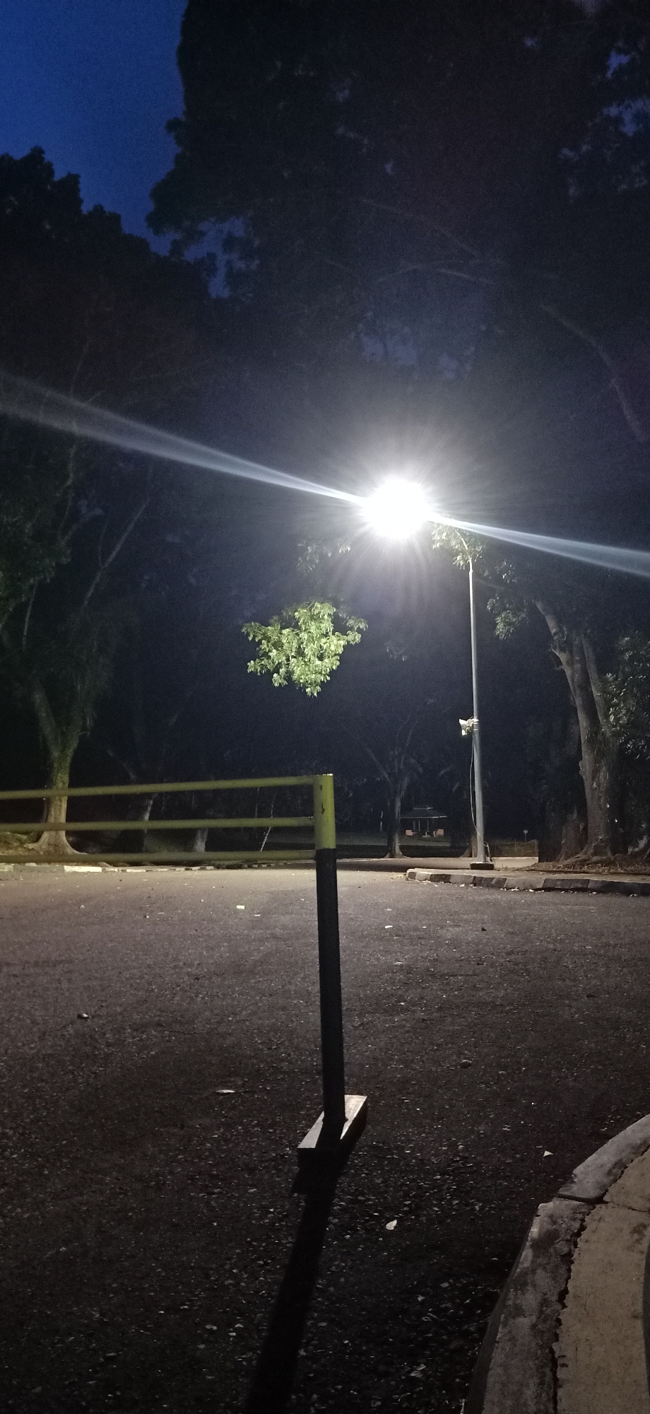 The image depicts a dimly lit outdoor scene at dusk or night, likely in Palohgadeng, Indonesia. In the foreground, a dark, rectangular post stands on the asphalt ground, casting a long shadow. To the left, a yellow horizontal barrier with vertical supports is visible. Beyond the barrier, trees with dense foliage are silhouetted against the darkening sky. A bright streetlamp illuminates the upper-middle portion of the image, creating a strong glare and a beam of light extending diagonally. The sky is a deep blue, indicating it is either early evening or late night. In the background, more trees and a faint structure, possibly a small building or shelter, are discernible. The ground is paved and appears to be a road or pathway. The overall atmosphere is quiet and still, characteristic of a park or public area after dark.