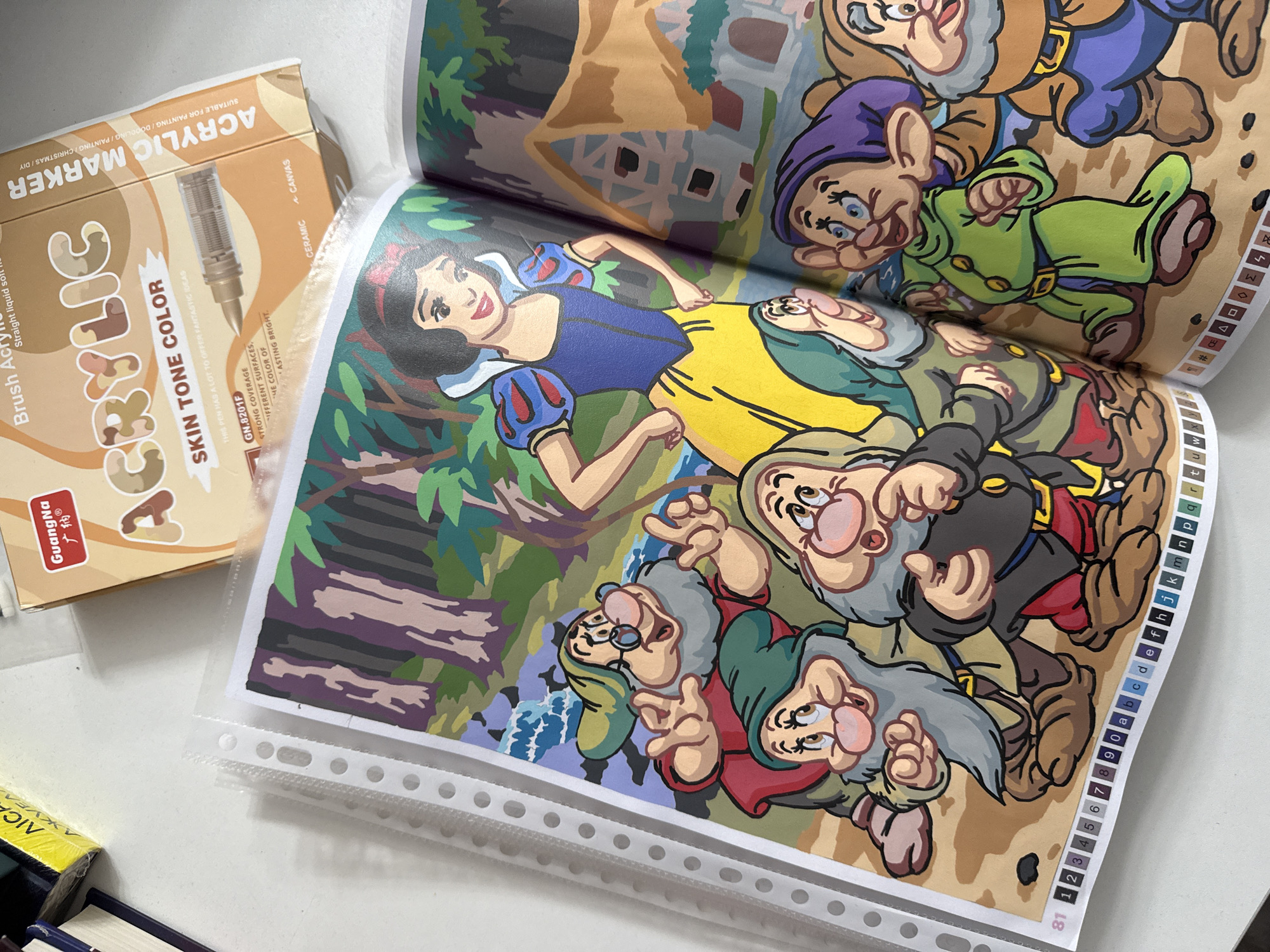 The image shows a close-up view of an open coloring book featuring Snow White and the Seven Dwarfs, with a box of "GuangNa Acrylic Marker Skin Tone Color" positioned behind it. The coloring book is placed on a white surface, likely a table, and is held open by a clear plastic protector with punched holes. The coloring book is open to a scene depicting Snow White with her signature blue and yellow dress, standing in a forest environment with the dwarfs gathered around her. The artwork appears to be a paint-by-numbers or similar coloring activity, with numbers and color swatches visible along the bottom edge of the left page. The box of markers is partially visible, with the brand name "GuangNa" and product details like "ACRYLIC MARKER" and "SKIN TONE COLOR" clearly legible. The overall scene suggests a crafting or hobbyist setting. There is no information in the image to indicate its location in the city of Łódź, Poland.