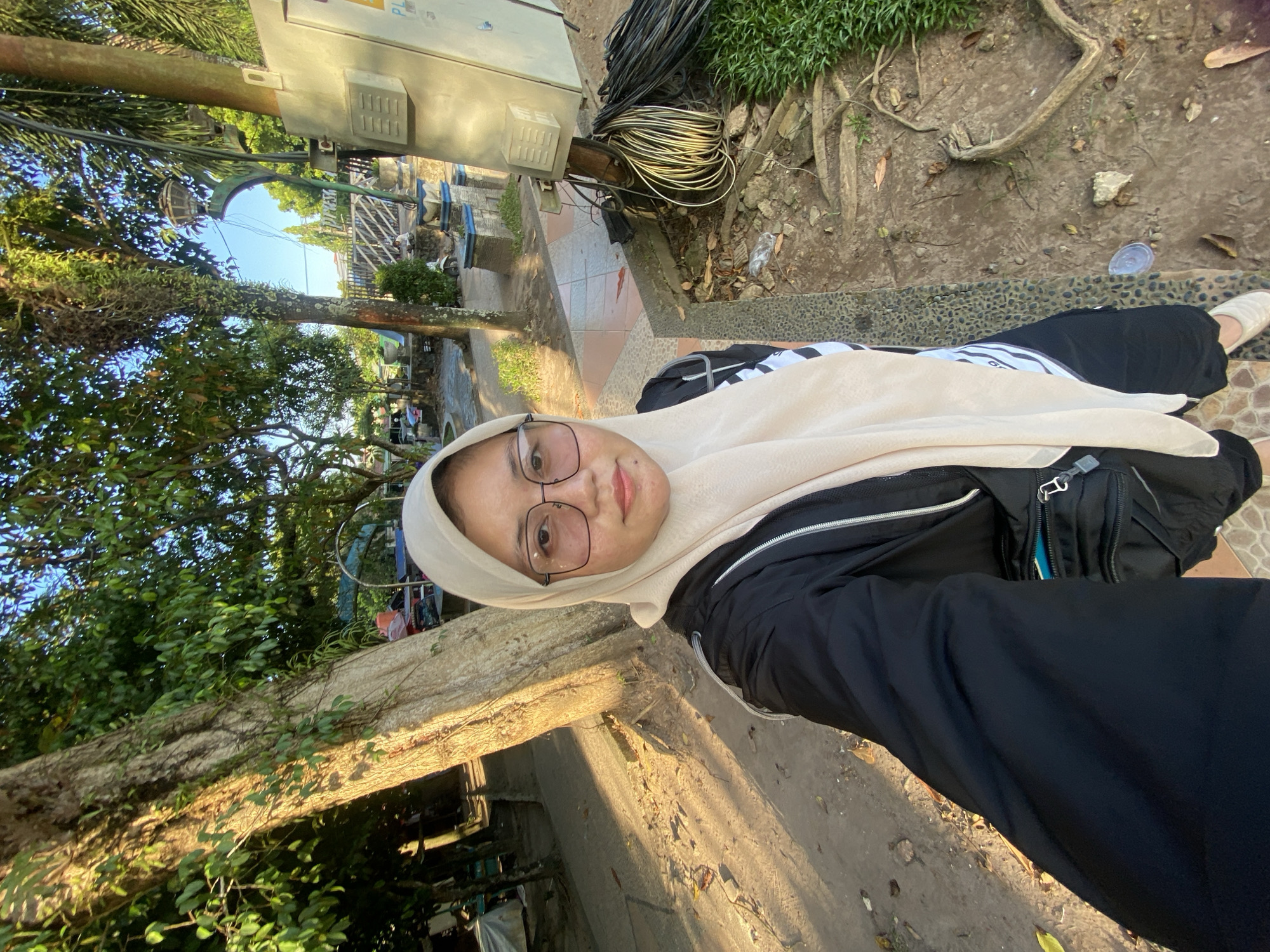 A woman wearing a cream-colored hijab and glasses takes a selfie outdoors. She is dressed in a black zip-up jacket over a striped shirt, and black pants. A black bag with a blue interior is slung across her body. The background shows a large tree trunk to her left, with dappled sunlight falling on the sandy ground. Beyond the tree, there are more trees and foliage, as well as some structures and what appear to be outdoor seating areas. To her right, there is a utility box with coiled wires and a patch of green grass. The scene suggests an outdoor public space, possibly a park or a roadside area, in Martapura, Indonesia.