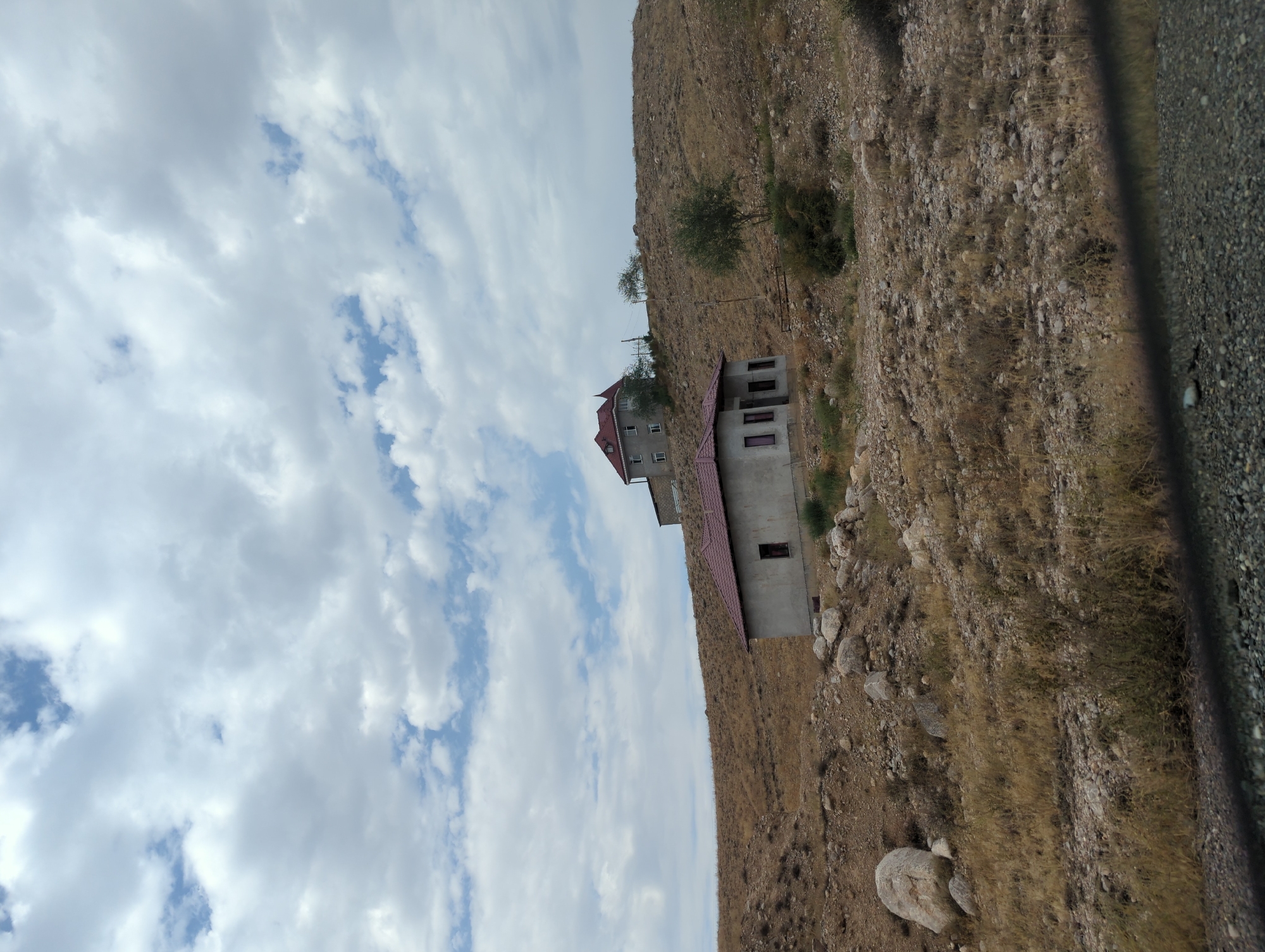 The image depicts a semi-arid, hilly landscape with two unfinished houses. The foreground shows dry, yellowish grass and scattered rocks. The lower house, closer to the viewer, is a single-story structure with a reddish-brown roof and visible windows. It appears to be under construction, with exposed concrete walls. Further up the hill, a larger, multi-story house with a similar reddish-brown roof is visible. This house also has multiple windows and an unfinished appearance. The terrain is uneven and sparsely vegetated with dry grasses and a few small trees. The sky is cloudy with patches of blue visible. The scene is located in the city of Samarkandyk, Kyrgyzstan, and shows rural or suburban development in a challenging environment. There are no people or animals visible in the image.