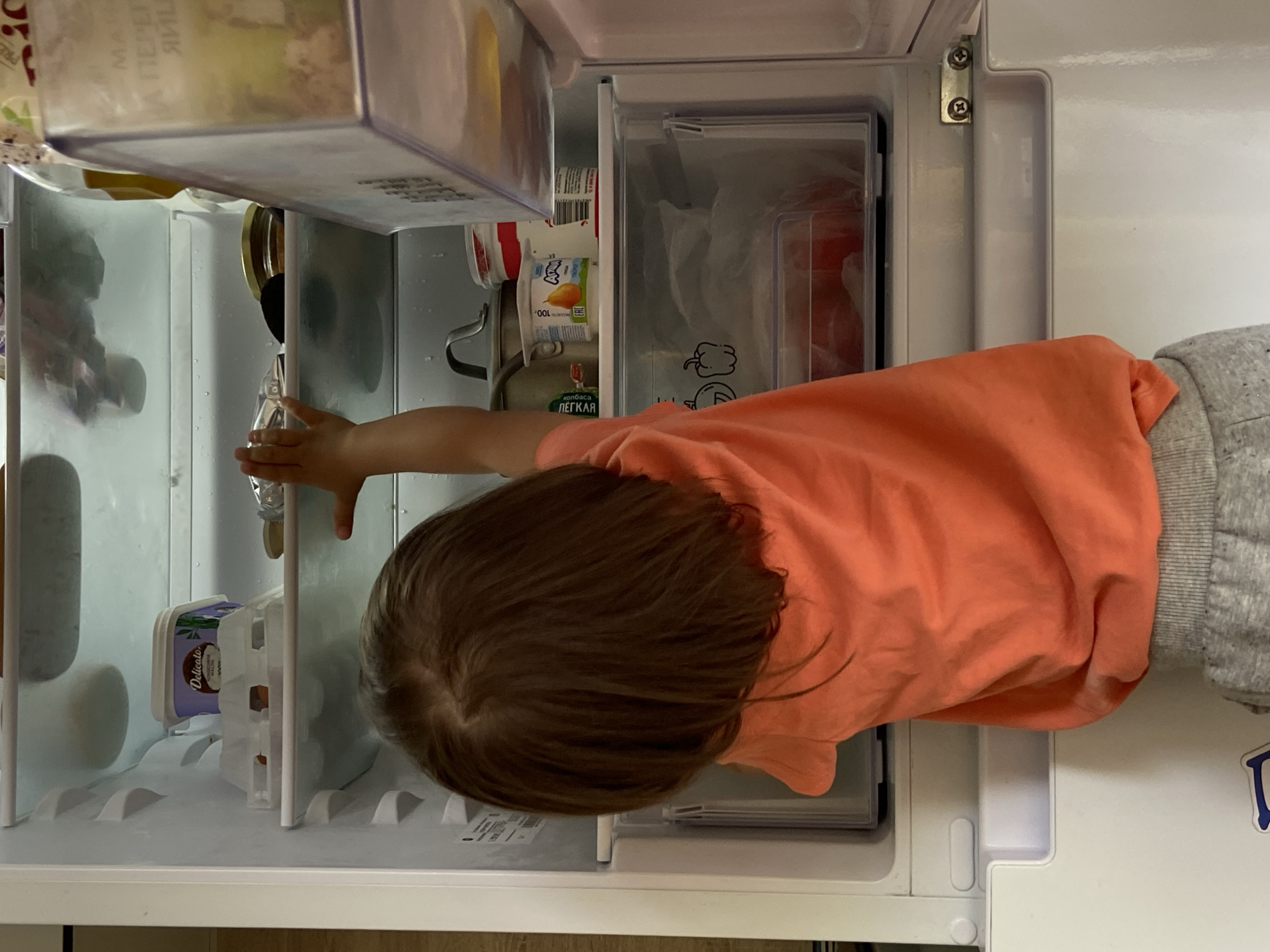 The image shows a young child in an orange t-shirt and grey sweatpants reaching into an open refrigerator. The child's hand is extended towards a clear plastic bottle with a white cap, which is sitting on a shelf. The refrigerator is well-stocked with various food items, including what appears to be yogurt containers and a jar with a red lid. In the background, there are multiple shelves and drawers within the refrigerator, some of which contain other food items and packaging. The overall scene suggests a domestic setting, possibly a kitchen, where the child is interacting with the refrigerator. The lighting in the image is bright, suggesting it's daytime. There is text visible on some of the items in the refrigerator, but it is mostly blurry and difficult to read. One label on a container clearly shows "Delicato" in purple lettering. Another label visible on a small white container has an illustration of a pear and says "100." A longer label on a green package reads "ХОЛБА" and "ЛЕГКАЯ." The image is oriented vertically, as if the camera was held upright.