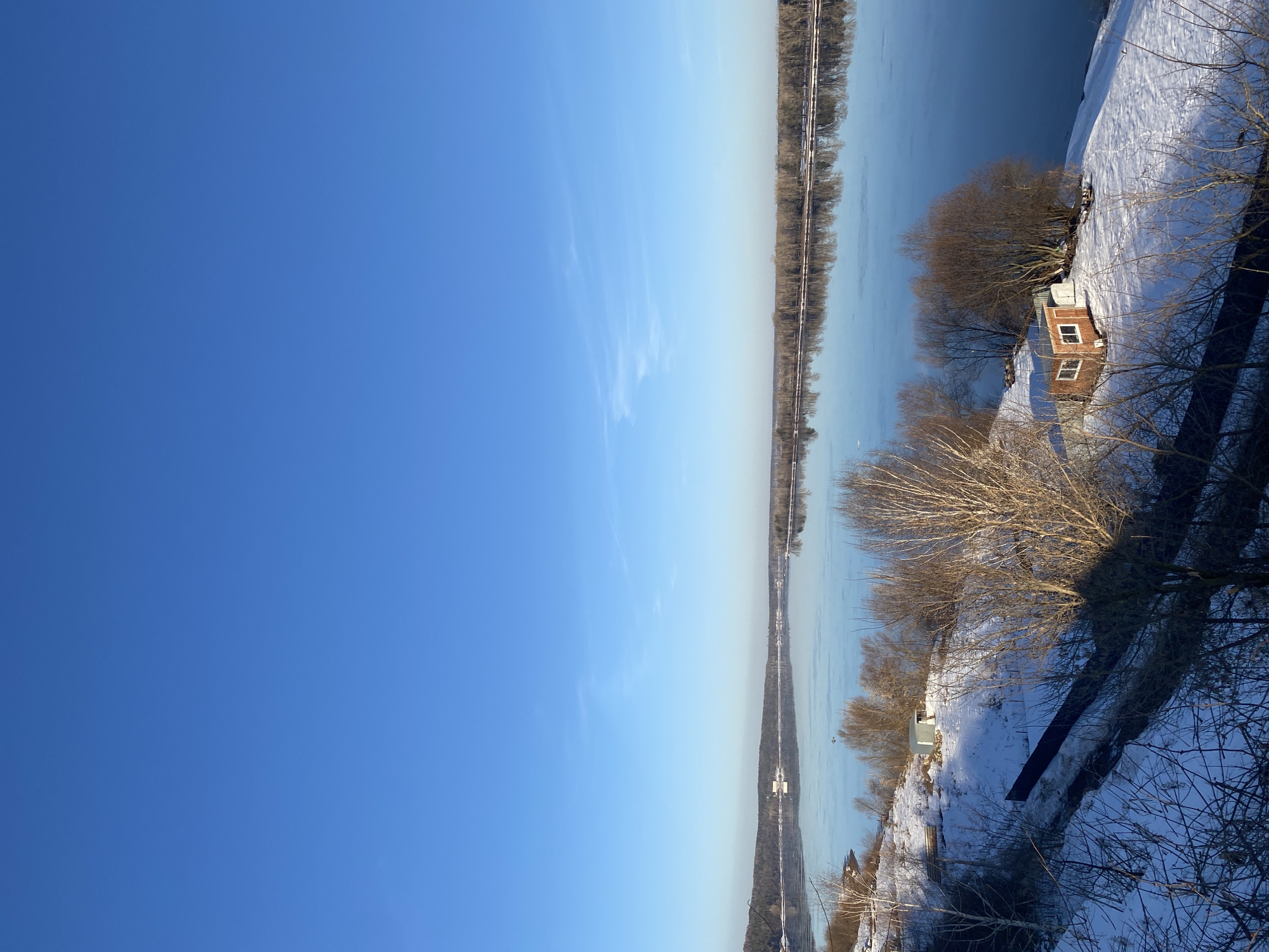 The image depicts a view of the Istra River in the city of Ольховка, Russia, during winter. The sky is clear and blue. The river is mostly frozen over. Along the riverbank, there are bare trees and patches of snow. A small brick building is visible near the shoreline.